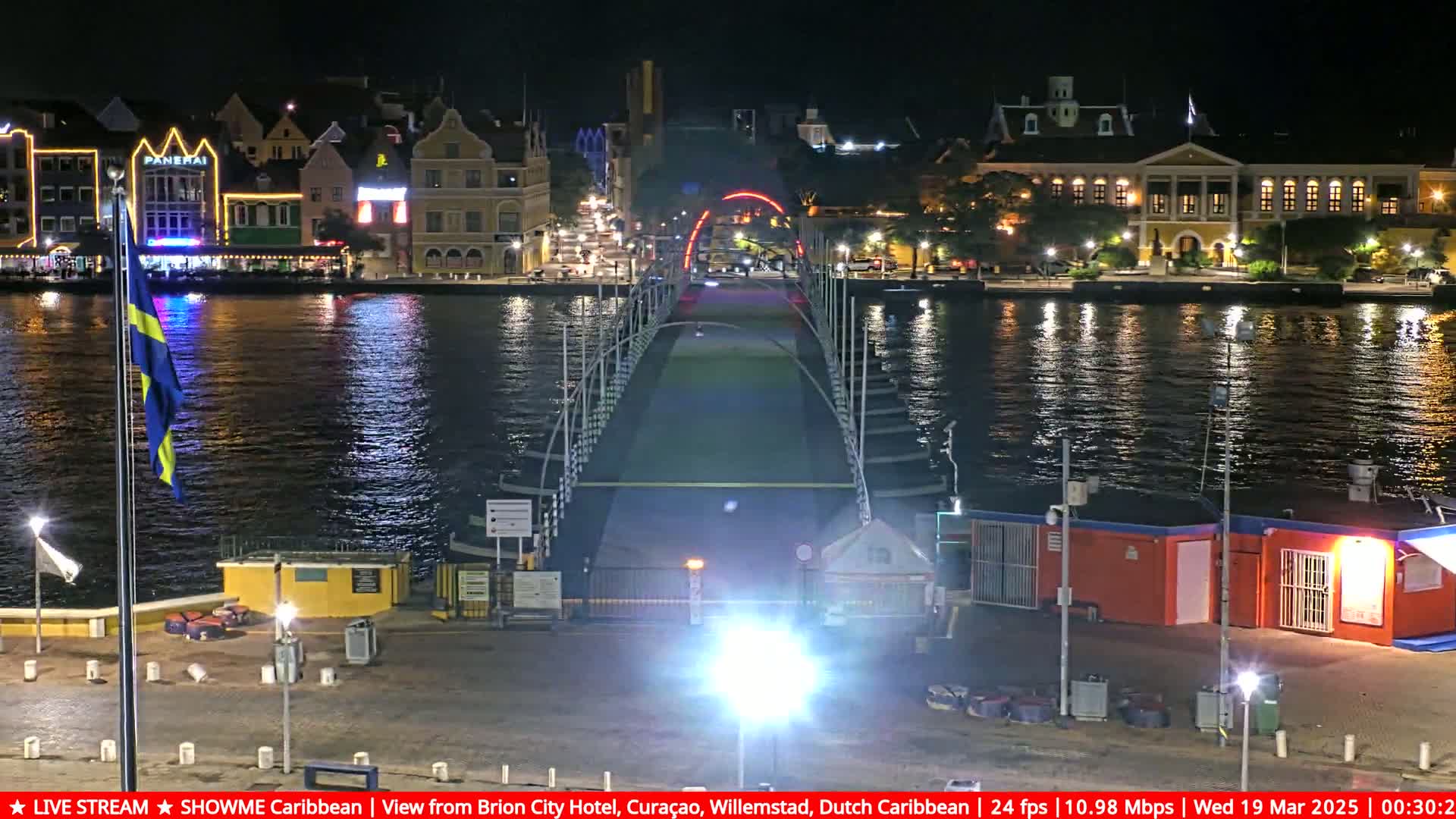 A nighttime view of a pontoon bridge leading to brightly lit buildings along a calm waterway, with a flagpole displaying a flag in the foreground.