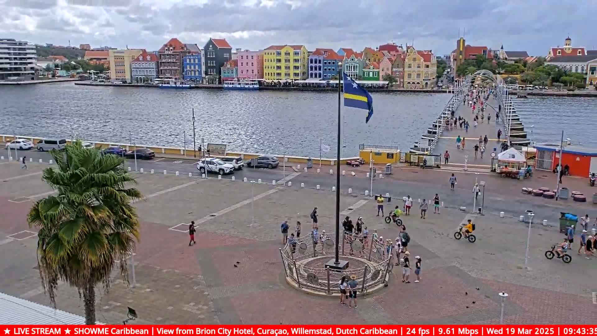 A waterfront plaza with a flagpole and a pontoon bridge, under an overcast sky, is filled with people and vehicles; colorful buildings line the opposite shore.