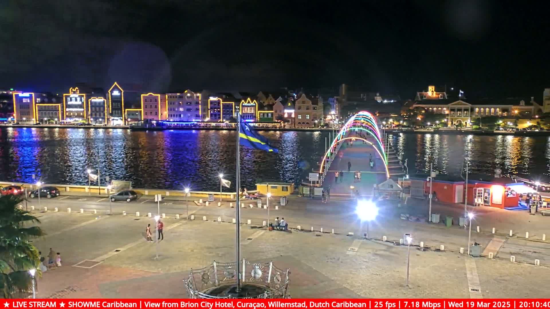 A nighttime view of a waterfront city, brightly lit buildings across a calm body of water, with a colorful illuminated bridge and a sparsely populated plaza in the foreground.