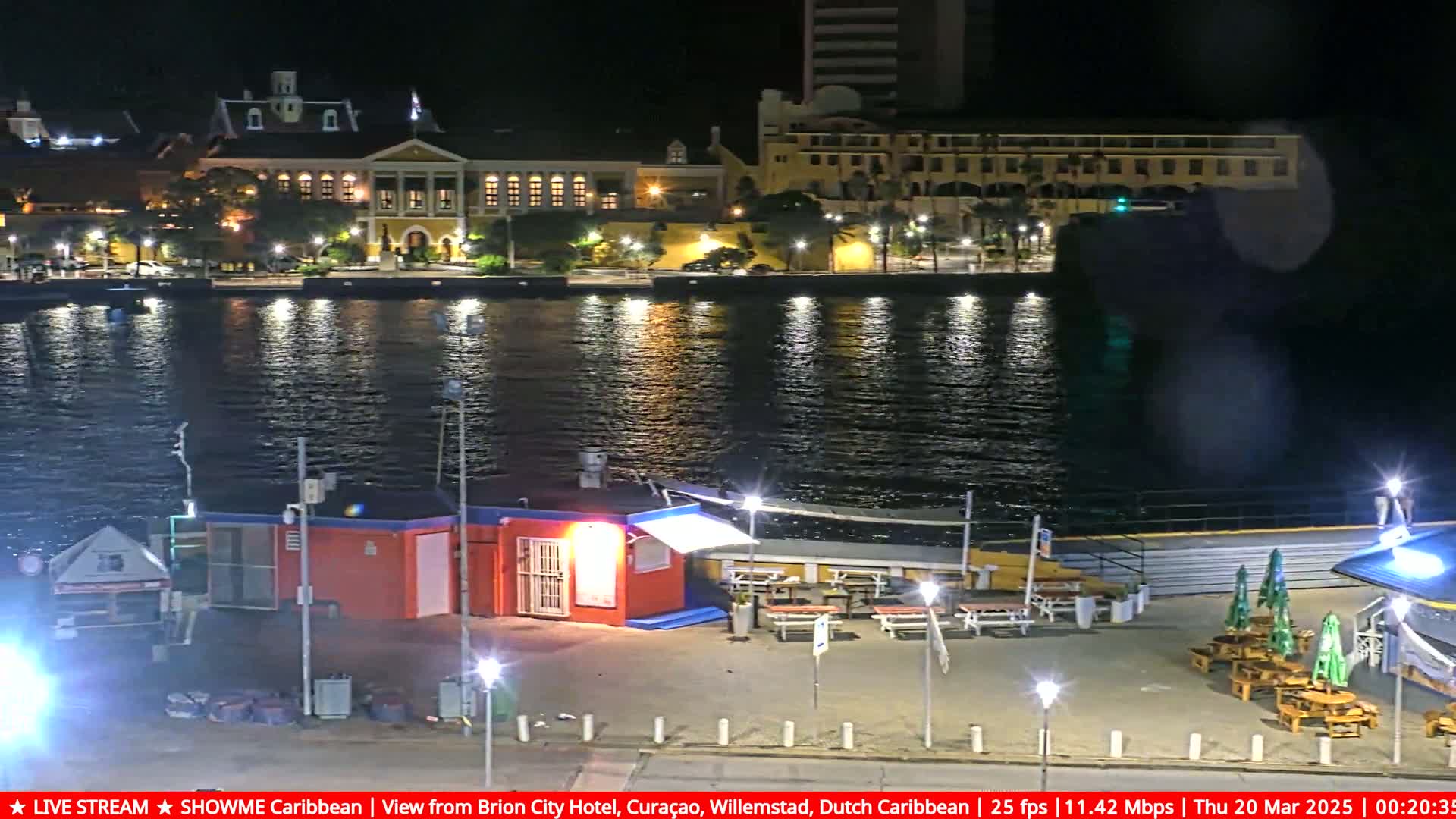 A nighttime view across calm water shows a waterfront area with illuminated buildings, small structures, and outdoor seating.