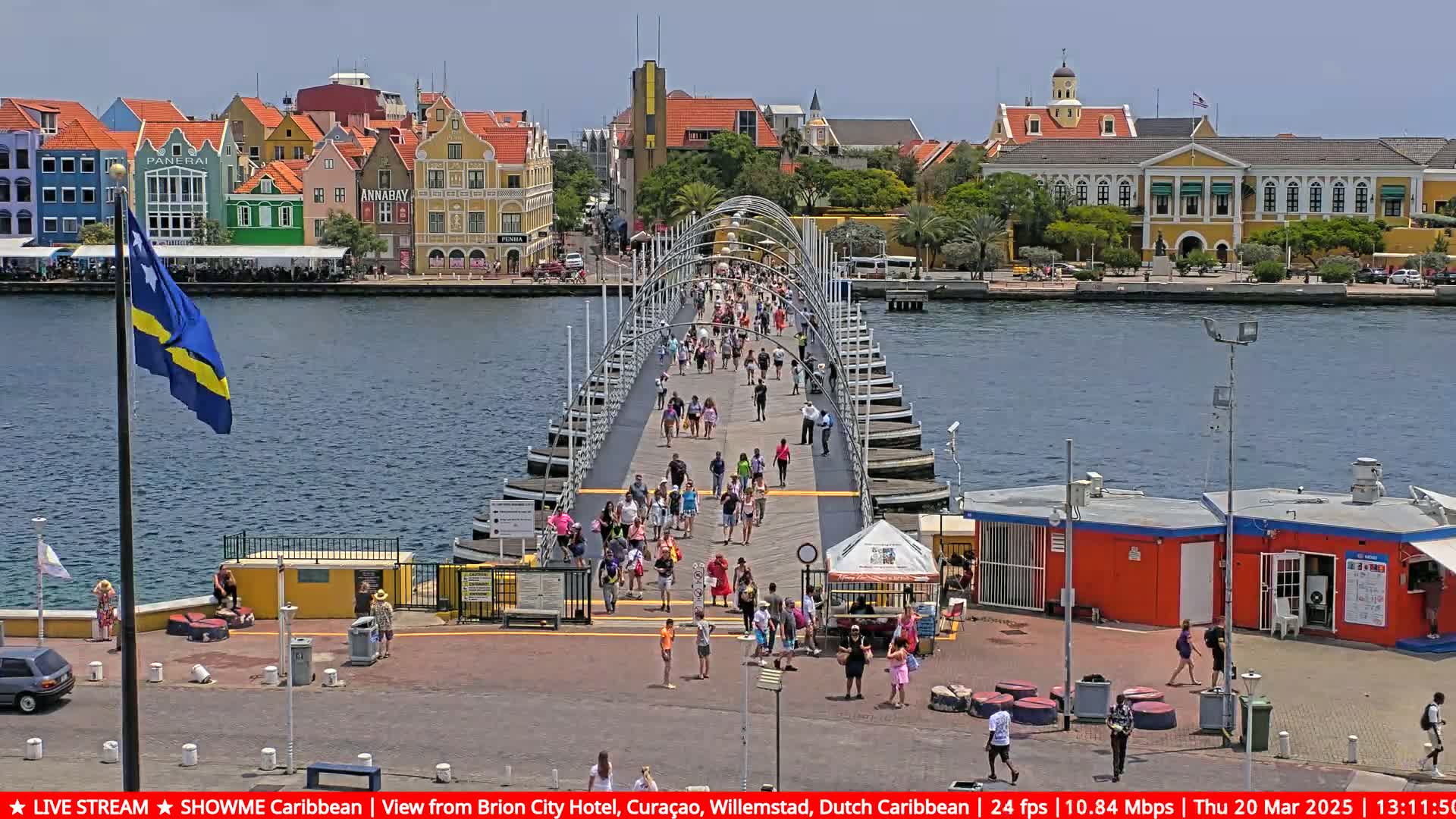 A pontoon bridge spans a waterway, with many people walking across it toward colorful buildings under a sunny sky.