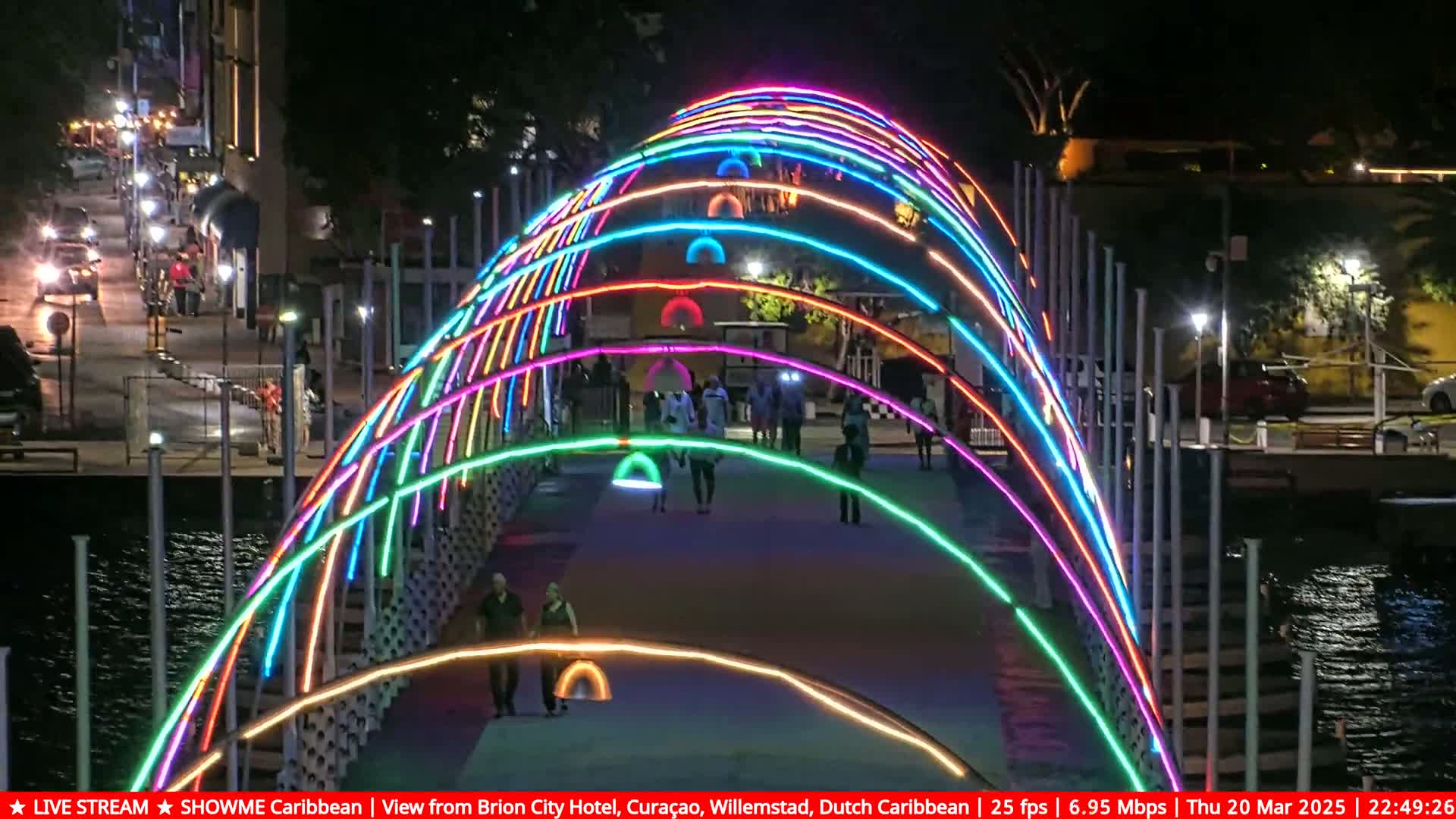 A pedestrian bridge at night, illuminated with multicolored neon lights, arches over a waterway with people walking along its path.