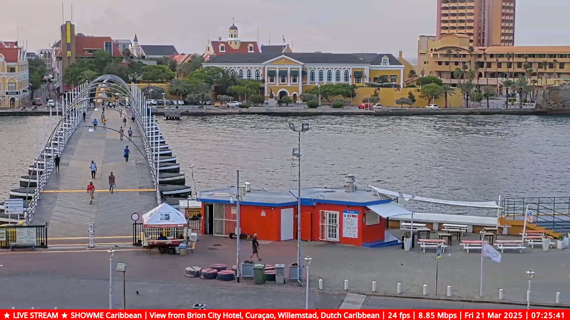 A pontoon bridge extends across a calm waterway toward a city with colonial-style buildings under an overcast sky.