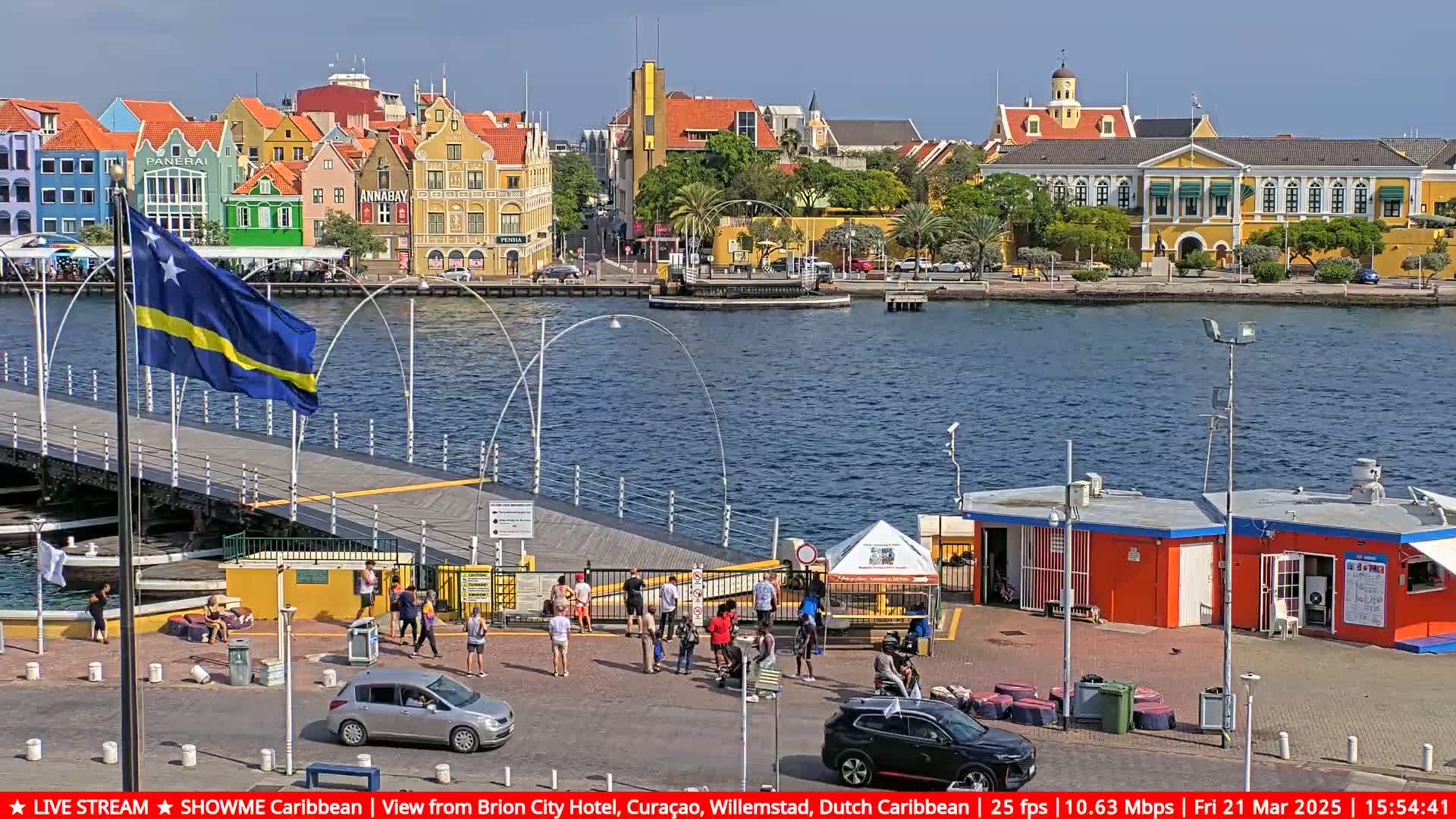 A Curaçao flag flies near a bridge over a calm body of water, with colorful buildings lining the shore under a sunny sky.