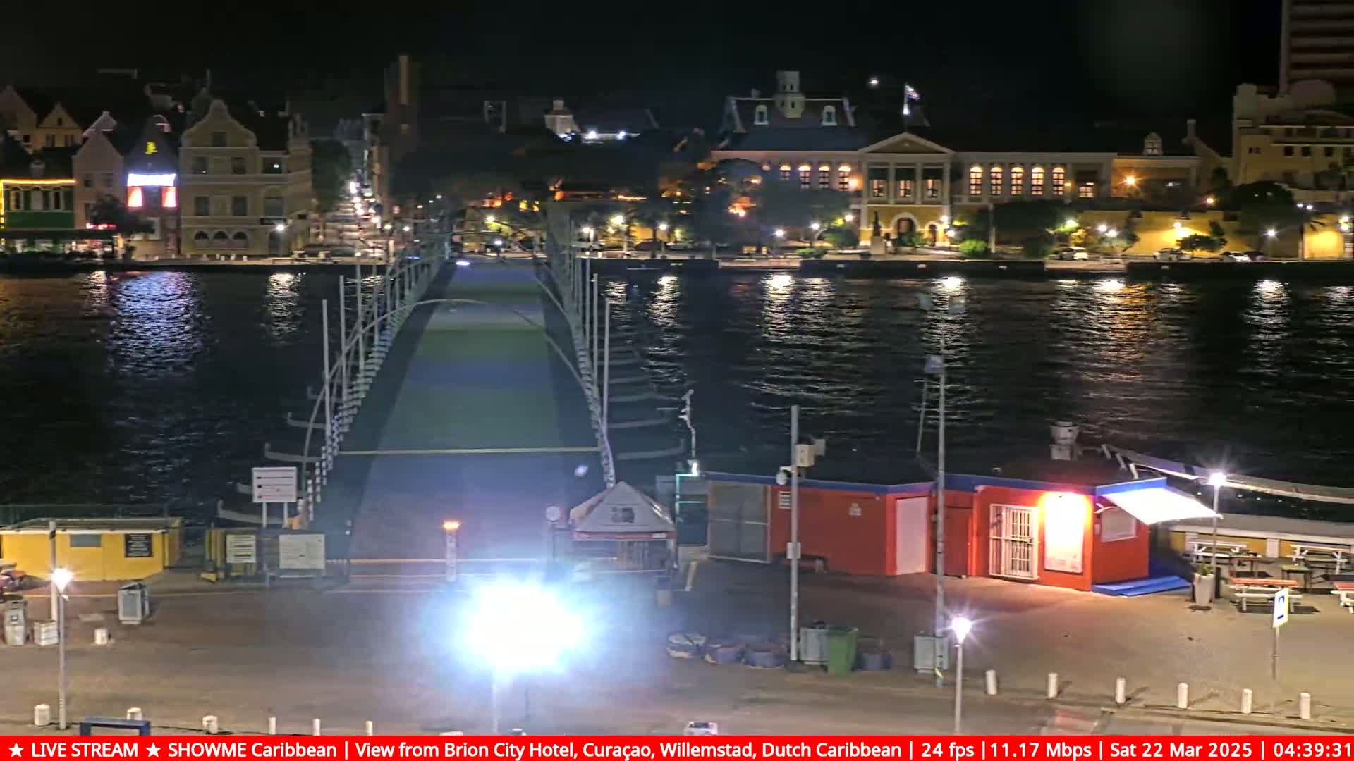 A nighttime view of a bridge spanning a calm body of water, with brightly lit buildings on the far shore.