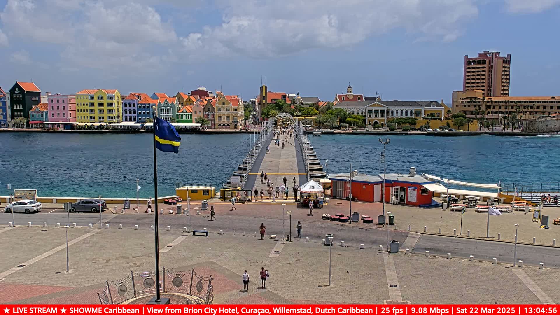 A long pier extends from a waterfront plaza toward colorful buildings across a calm blue body of water under a partly cloudy sky.