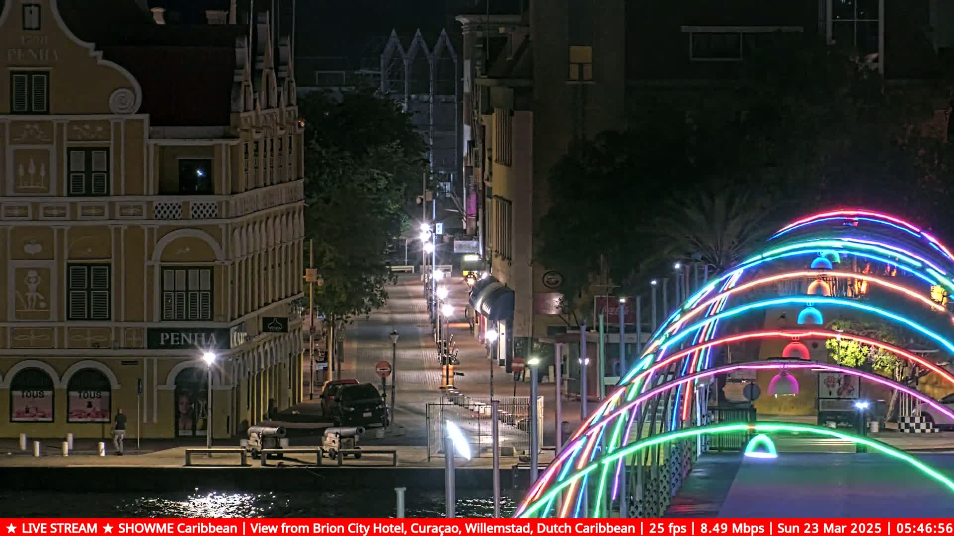 A nighttime view of a city street with colorful illuminated arches over a bridge, and buildings along the waterfront.