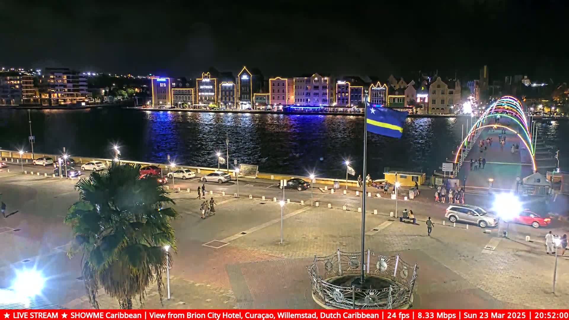 A nighttime view of a waterfront city with brightly lit buildings, a pedestrian bridge with colorful lights, and cars parked along a paved area.