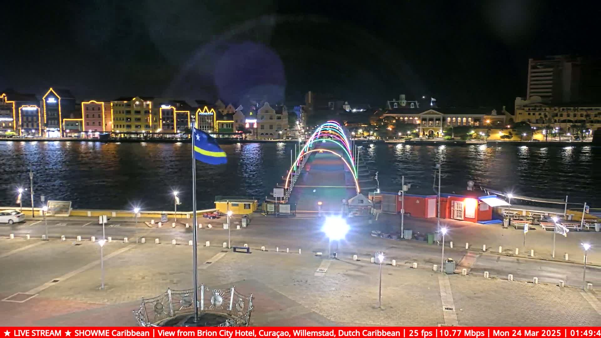 A brightly lit pedestrian bridge with rainbow-colored arches spans a waterway at night, with illuminated buildings lining the opposite shore.