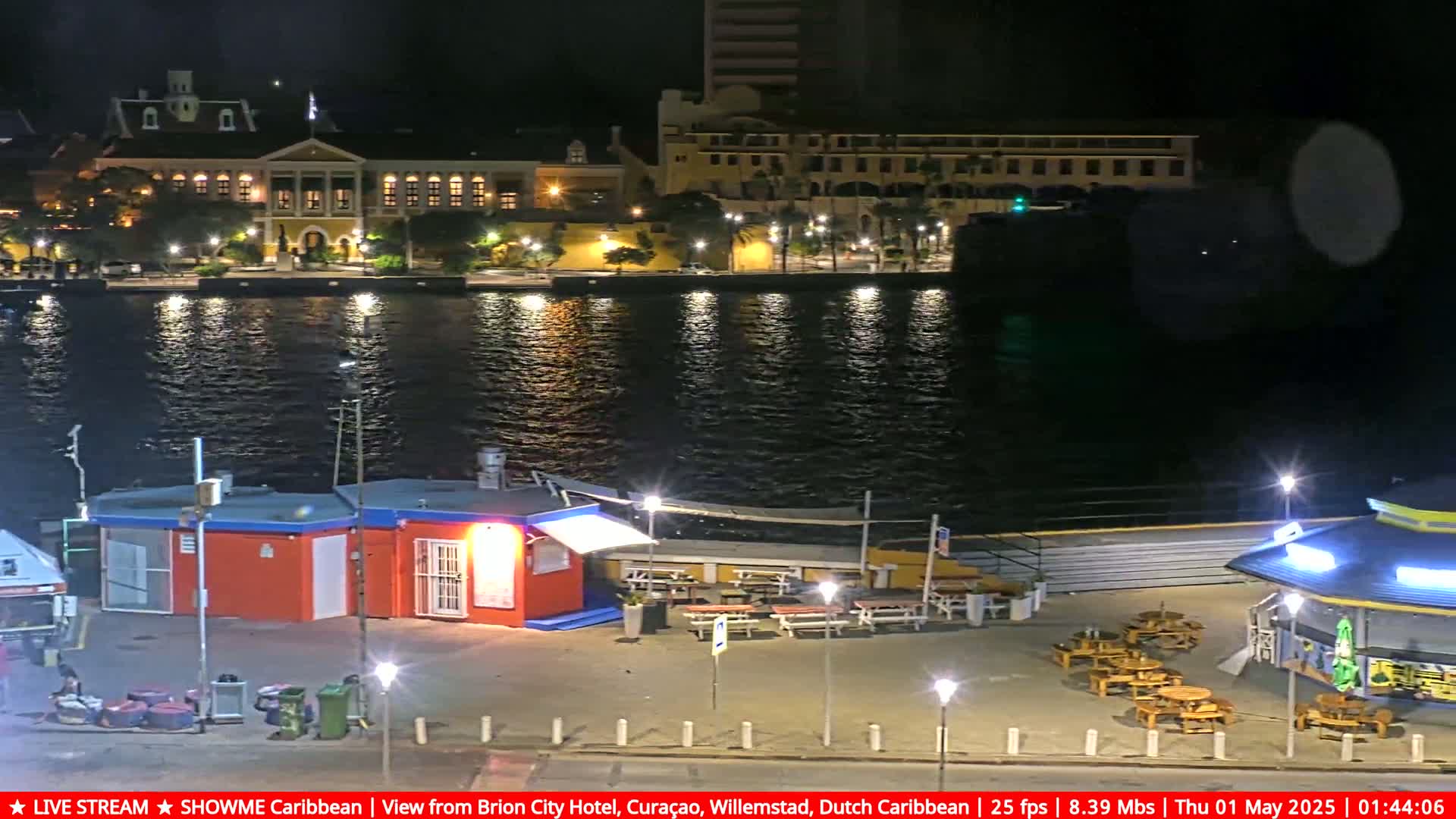 A nighttime waterfront scene shows illuminated buildings across a calm body of water, with outdoor seating areas and small structures along the shore.