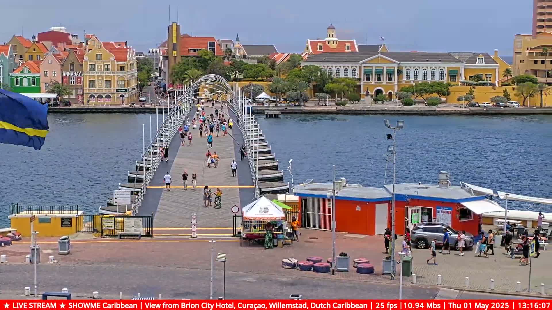 A pontoon bridge extends across a waterway, connecting to colorful buildings under a sunny sky; many people are walking across the bridge.