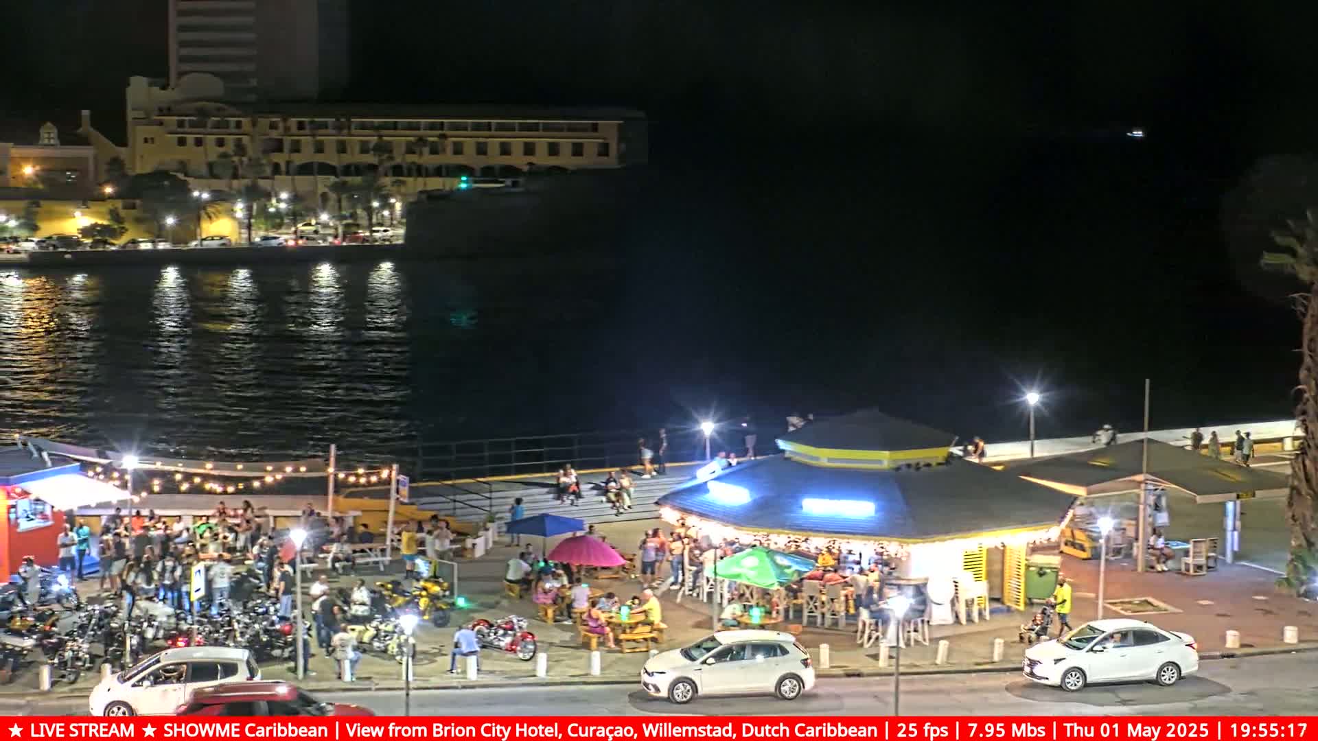 A nighttime waterfront scene shows a crowd of people gathered around an outdoor bar and restaurant next to a body of water with buildings and parked cars visible in the background.