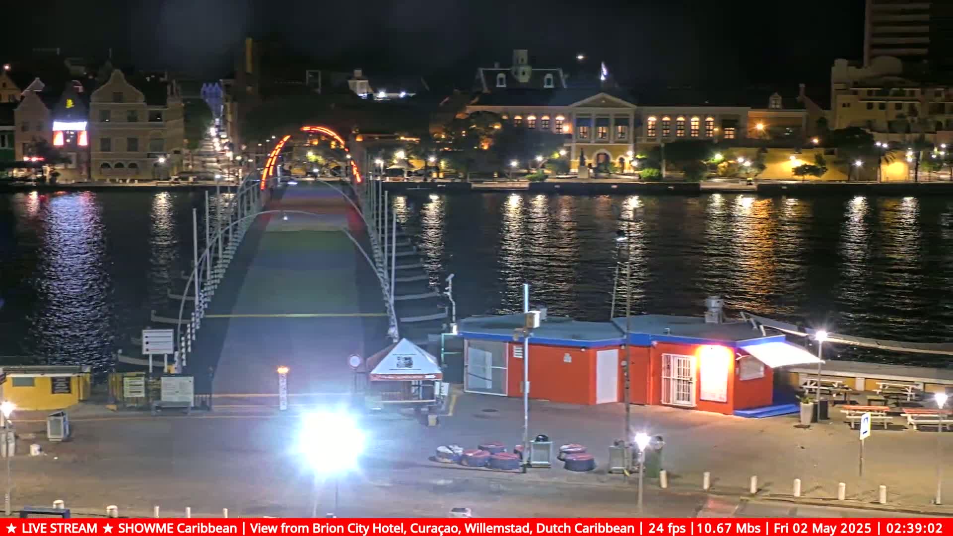 A brightly lit drawbridge spans a calm, dark waterway at night, with illuminated buildings lining the opposite shore.