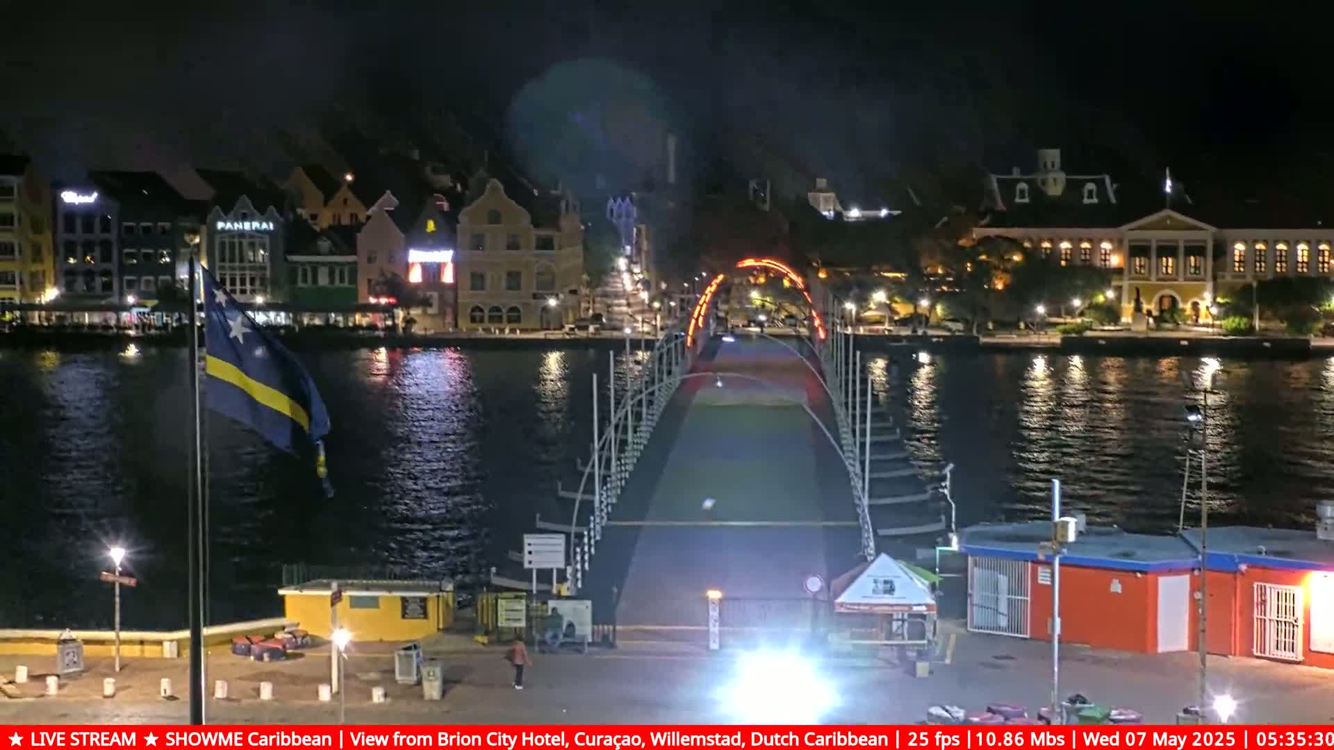 A nighttime view of a brightly lit bridge over a calm waterway, with colorful buildings lining the shore and a flag waving gently in the breeze.