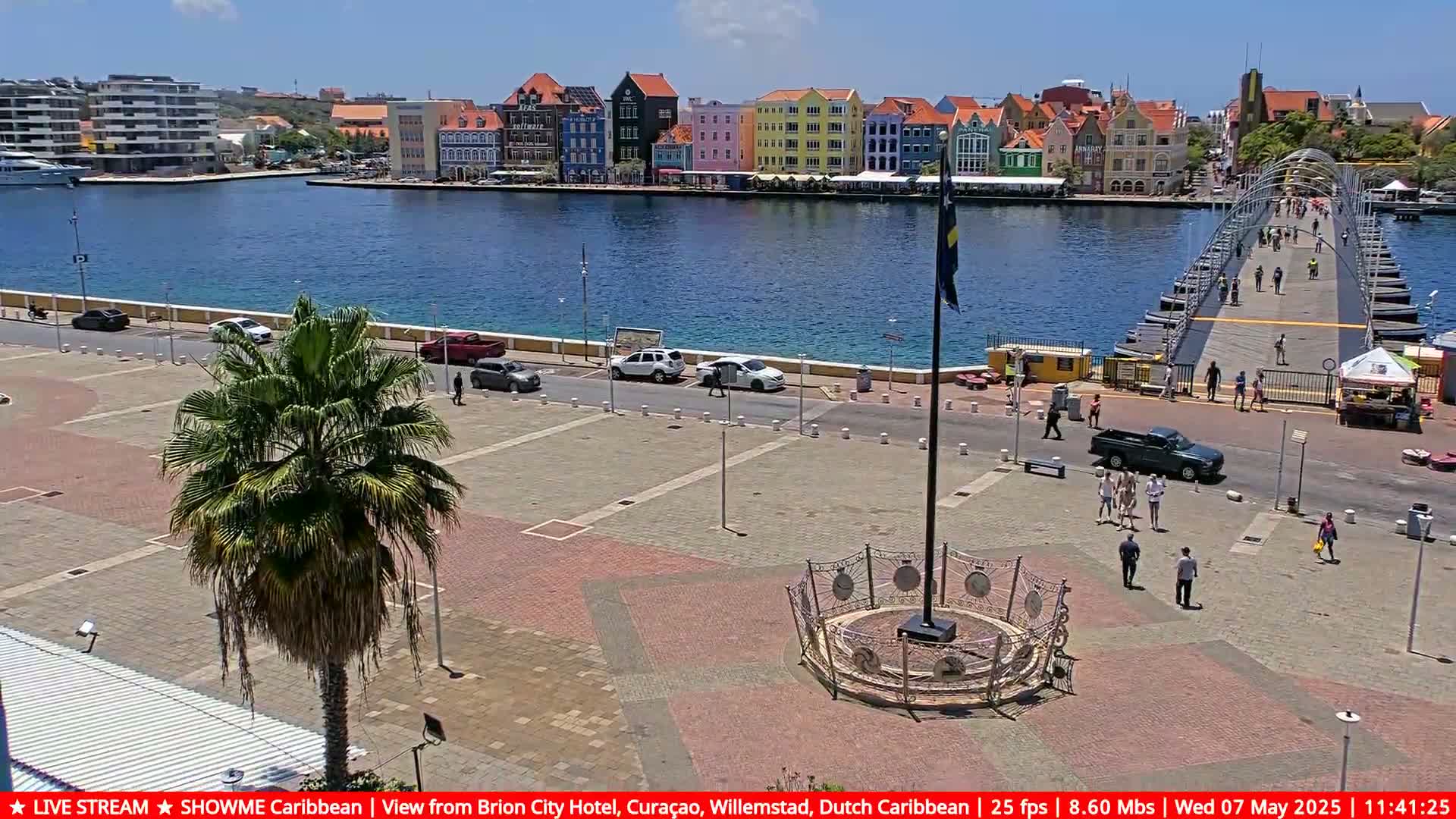 A sunny day reveals a waterfront plaza with a palm tree, parked cars, and pedestrians near a pontoon bridge leading to colorful buildings across a calm body of water.