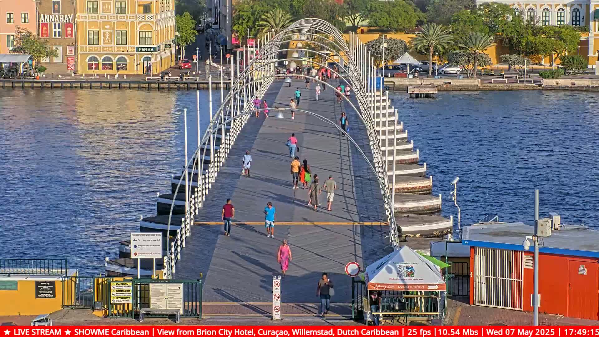 A pontoon bridge stretches across a waterway, with pedestrians walking along its path under a sunny sky; buildings line the waterway's edges.