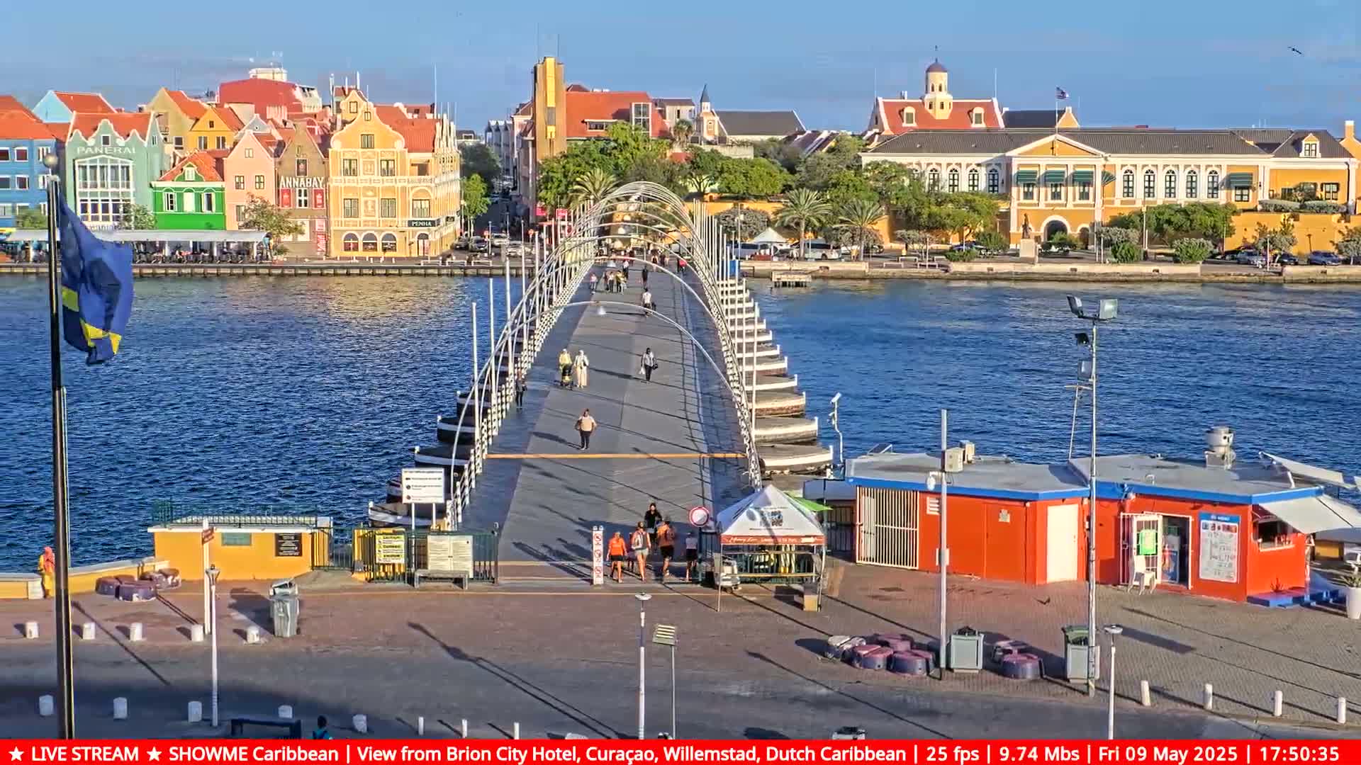 A pontoon bridge stretches across a calm body of water, connecting to colorful buildings under a sunny sky.