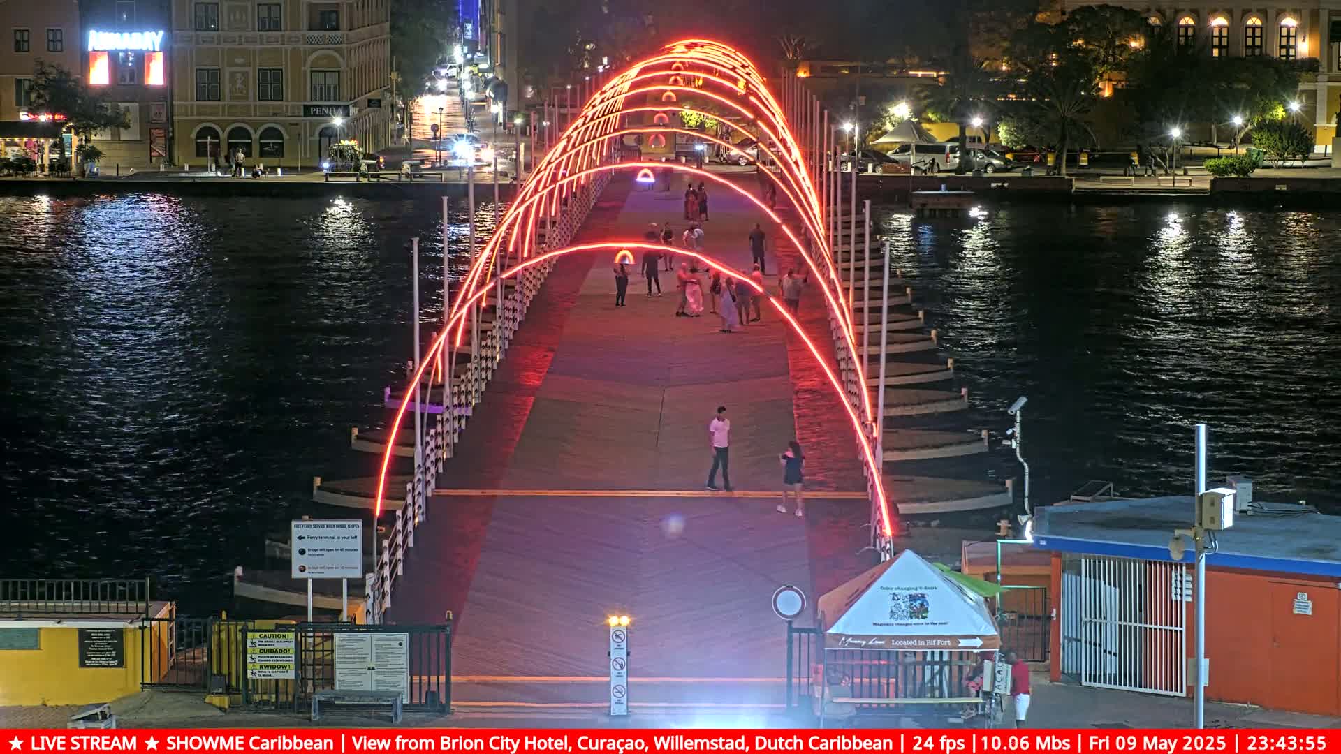A pontoon bridge illuminated with red lights at night spans a dark body of water, with a few people walking across it.