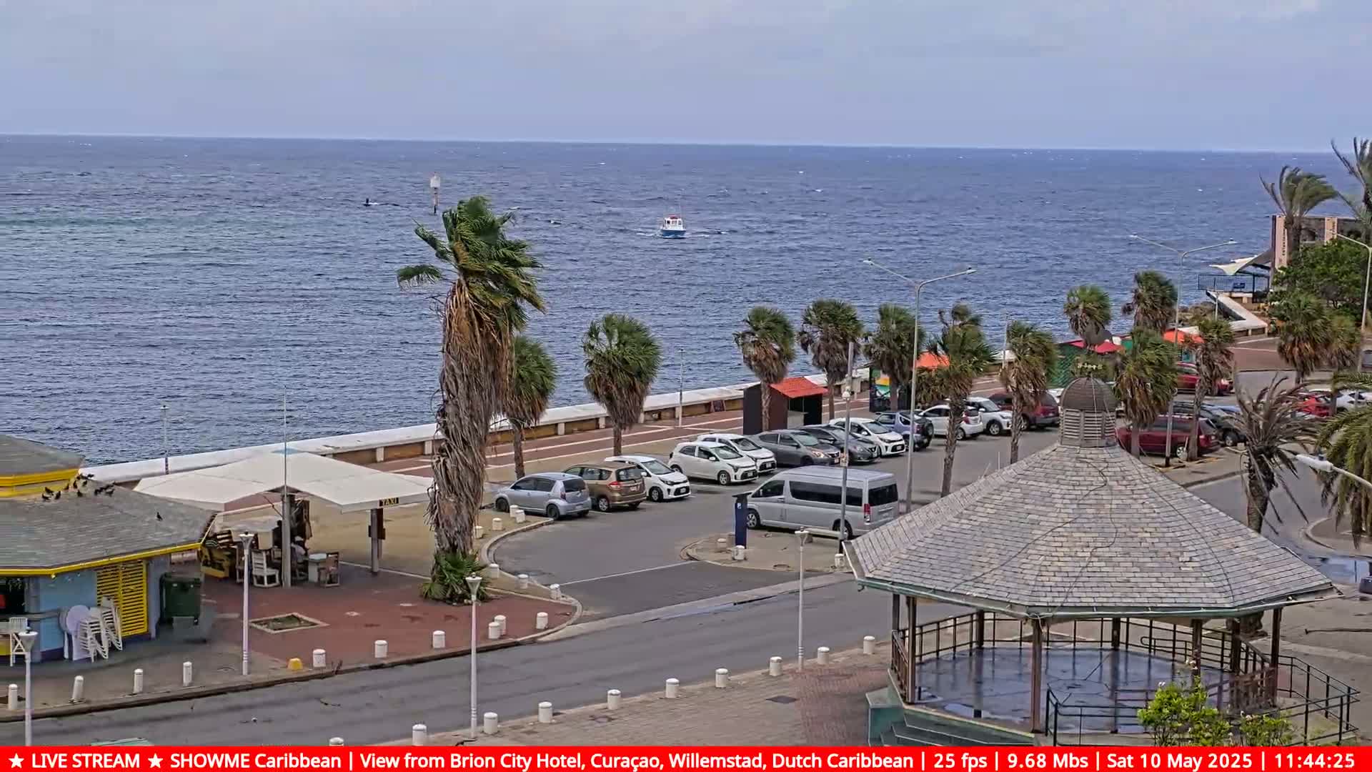 A parking lot with several cars is situated next to a waterfront promenade lined with palm trees, overlooking a calm ocean with a small boat visible in the distance, under a partly cloudy sky.