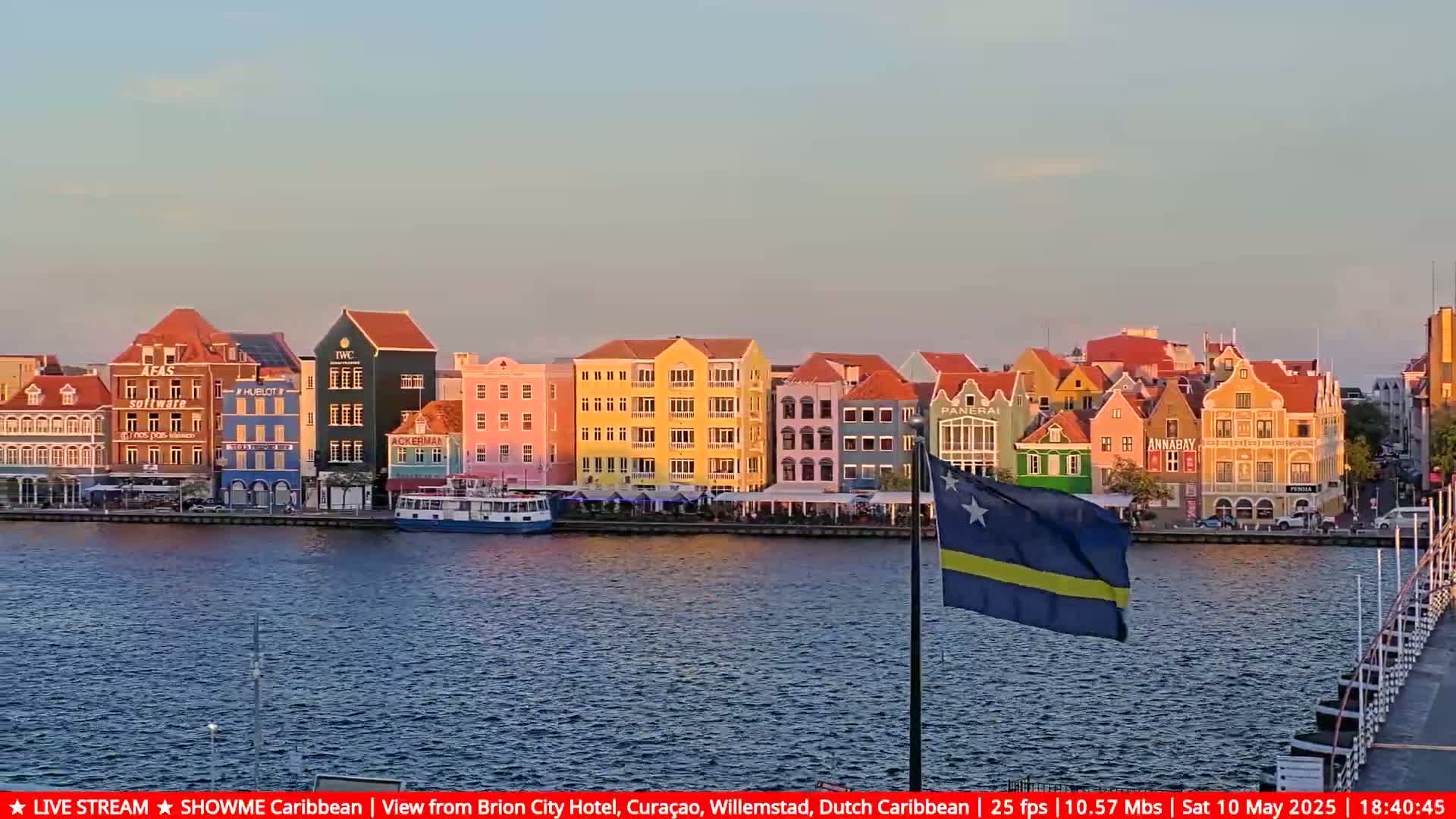 A row of colorful buildings lines a waterfront, with a Curaçao flag waving in the calm evening water.