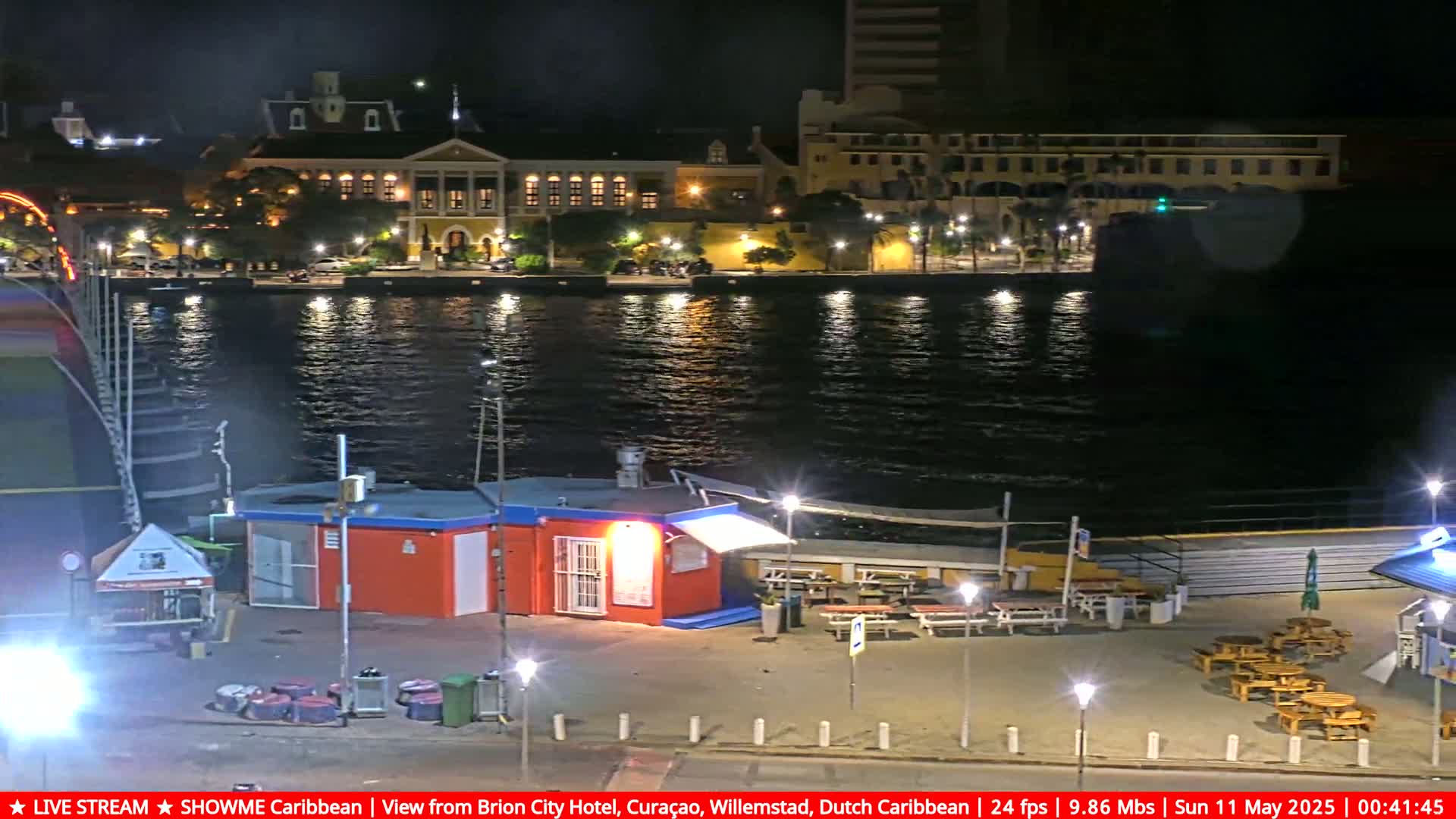 A waterfront scene at night shows a calm body of water reflecting lights from buildings across the water and a small outdoor eating area with several picnic tables.