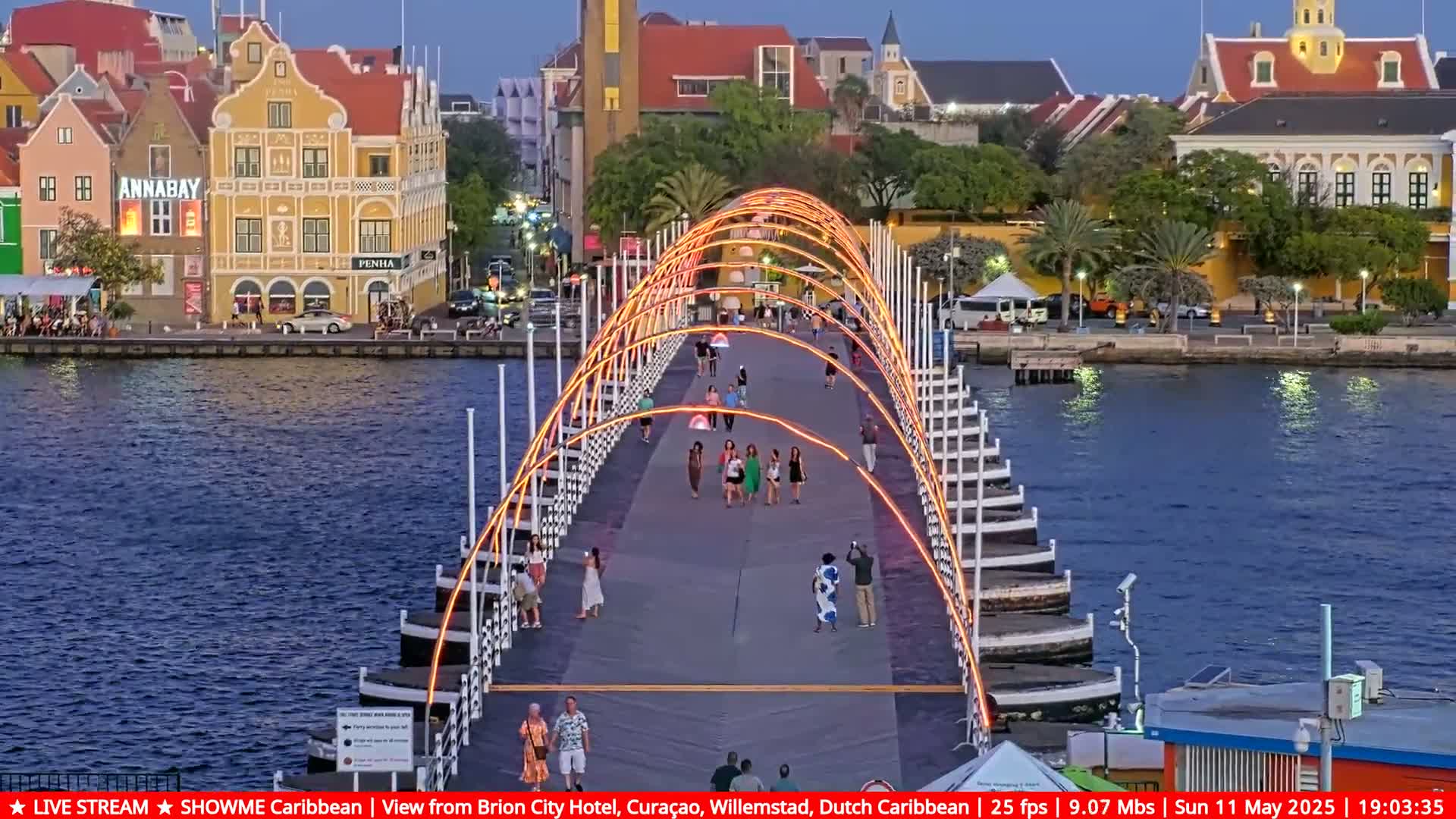 A brightly lit pedestrian bridge spans a waterway at dusk, with people walking across it and colorful buildings lining the shore.