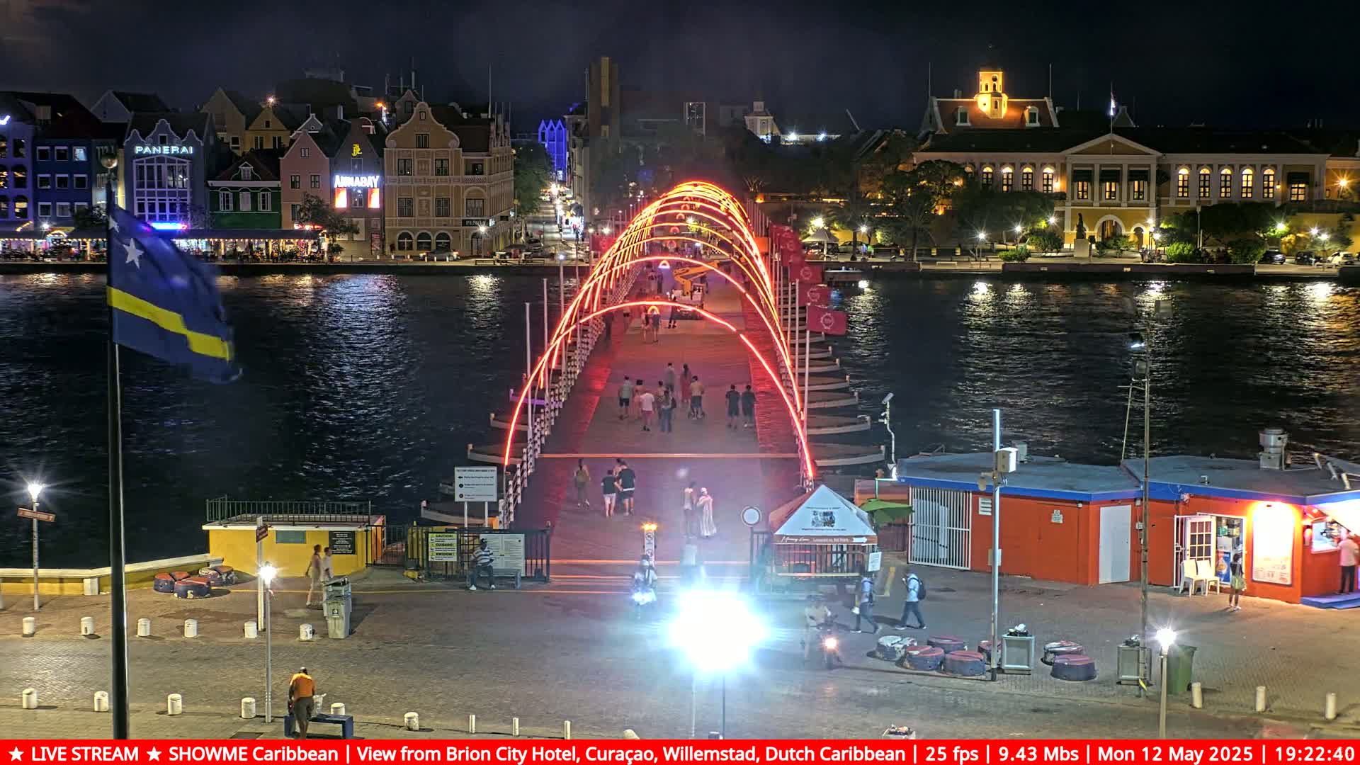 A brightly lit pedestrian bridge at night spans a waterway, with colorful buildings lining the shore.