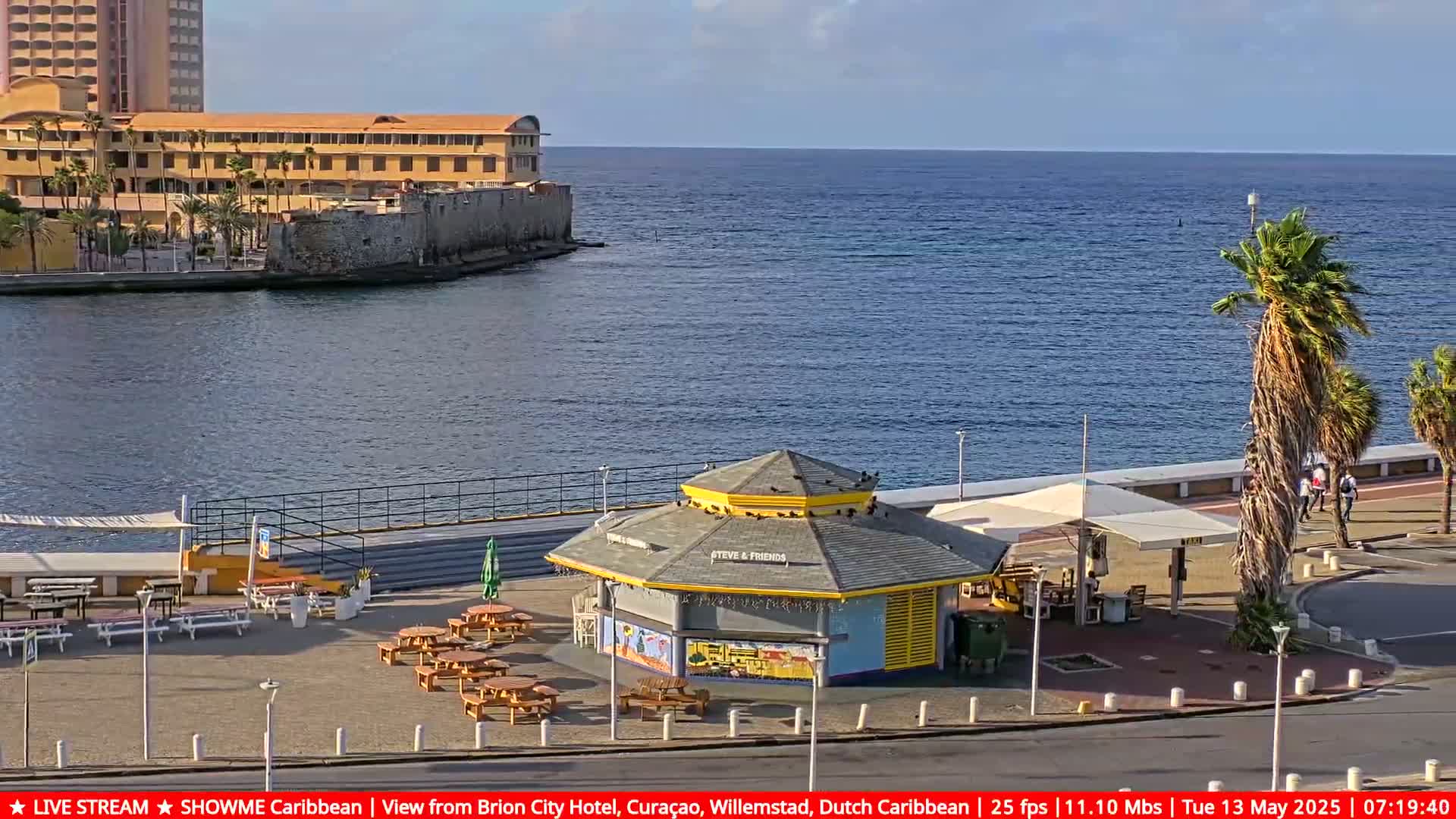 A waterfront scene shows a yellow kiosk with outdoor seating near a palm tree-lined promenade, a calm ocean, and a large building in the background under a clear sky.