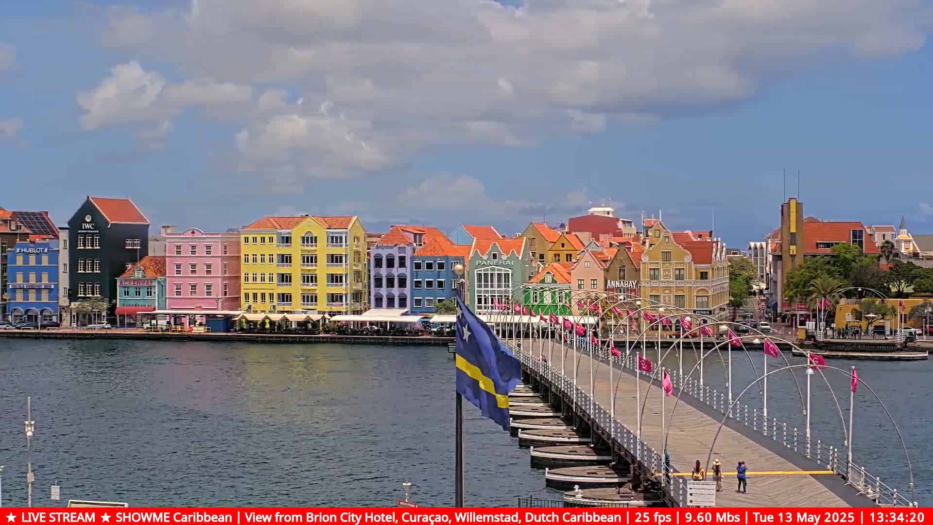 A pontoon bridge stretches across a calm waterway toward a waterfront lined with brightly colored buildings under a partly cloudy sky.