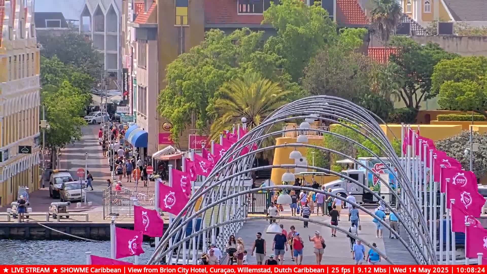 A sunny day shows a pedestrian bridge with pink flags over a waterfront street lined with colorful buildings and people walking.