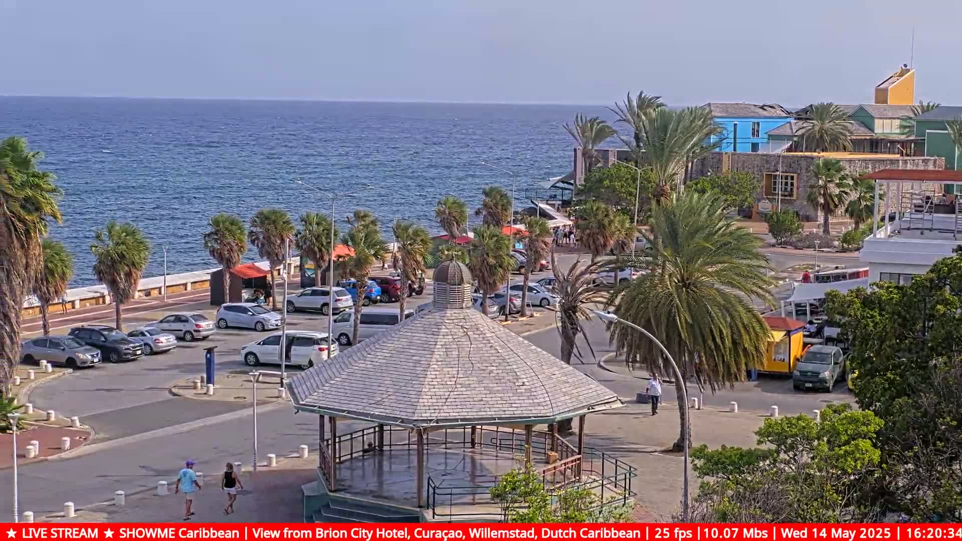 A seaside parking lot with a gazebo in the center, palm trees, and colorful buildings, under a sunny sky.