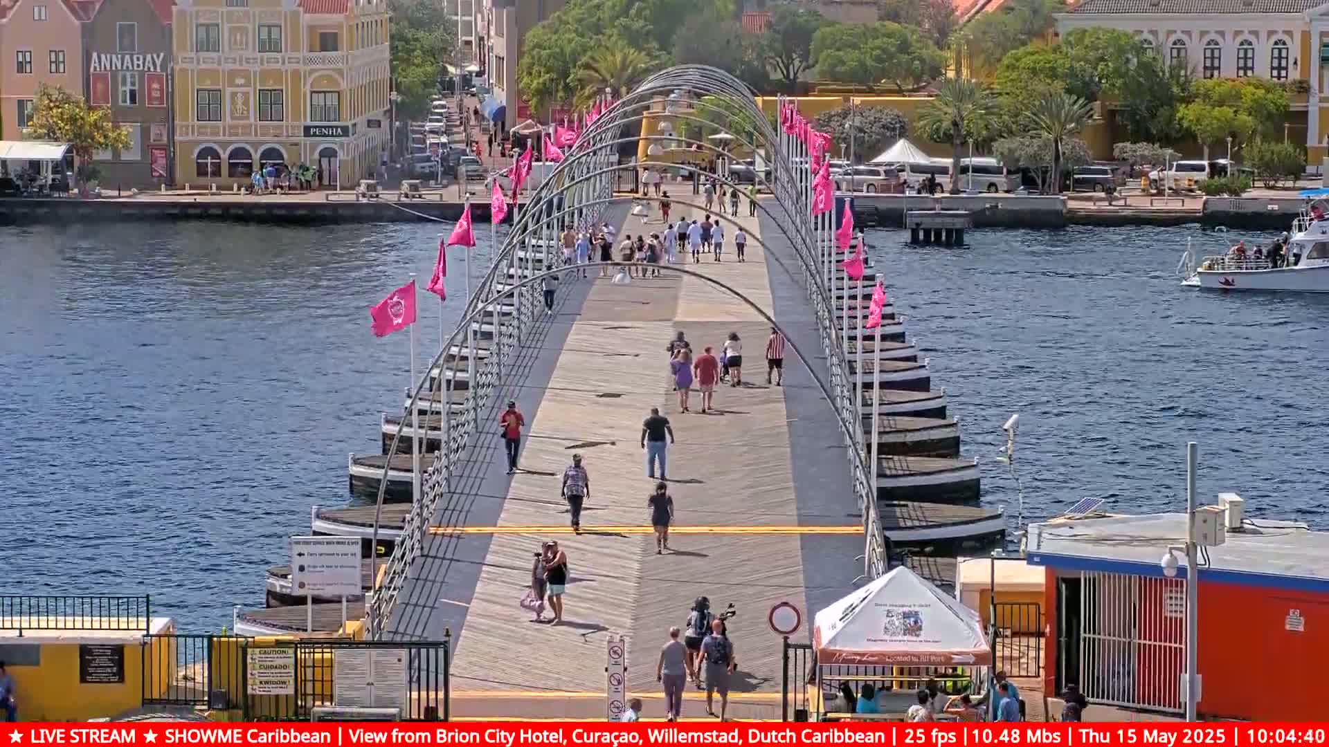 A pontoon bridge with people walking across it stretches over calm blue water, under a sunny sky, with colorful buildings visible in the background.
