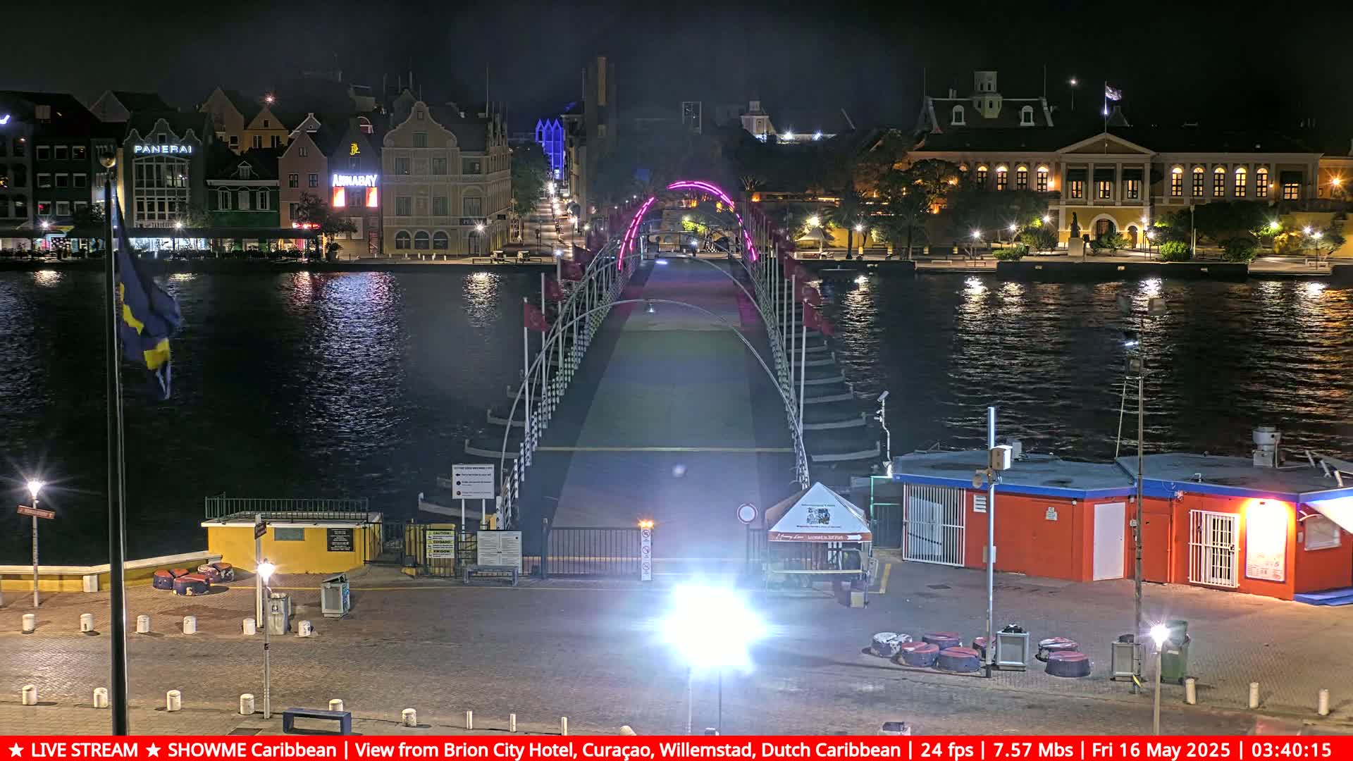A nighttime view of a pontoon bridge illuminated with pink lights spanning a calm body of water, with buildings lining the waterfront on either side.