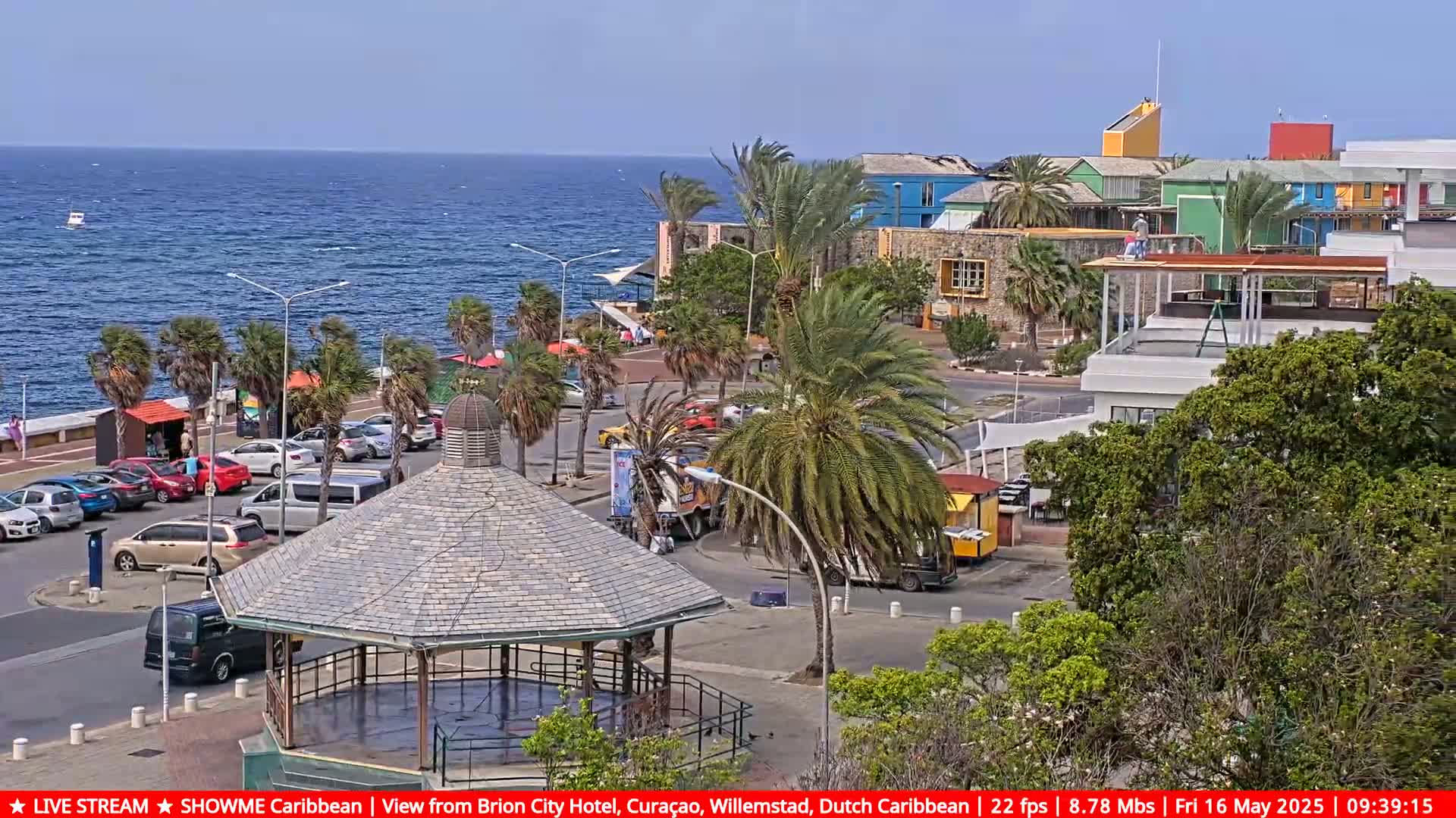 A seaside town square with a gazebo, palm trees, parked cars, and colorful buildings under a sunny sky.