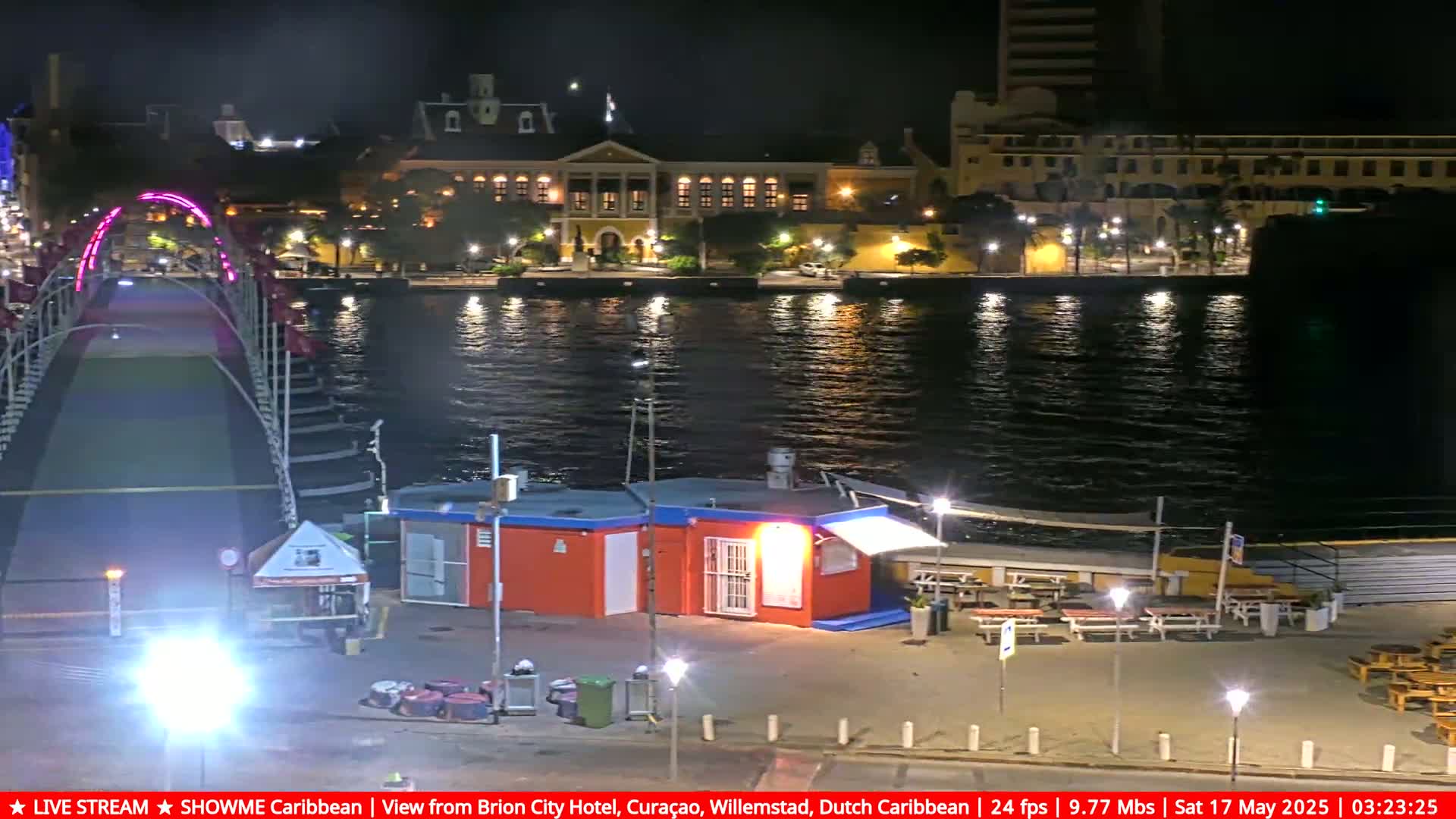 A nighttime view of a waterfront scene showing a lighted pontoon bridge, a building with outdoor seating, and a city skyline across a calm body of water.