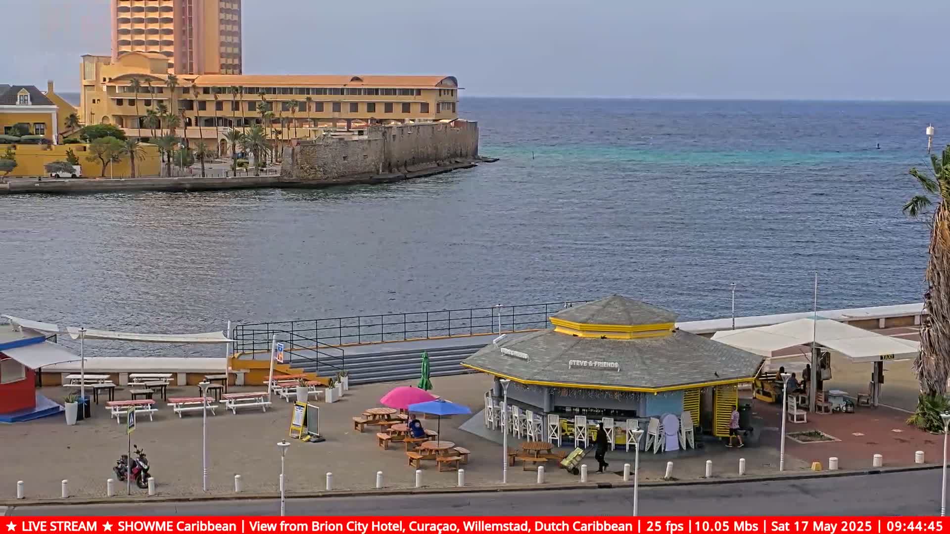 A waterfront scene shows a yellow building with outdoor seating near a calm ocean under a mostly clear sky, with a tall hotel visible in the background.