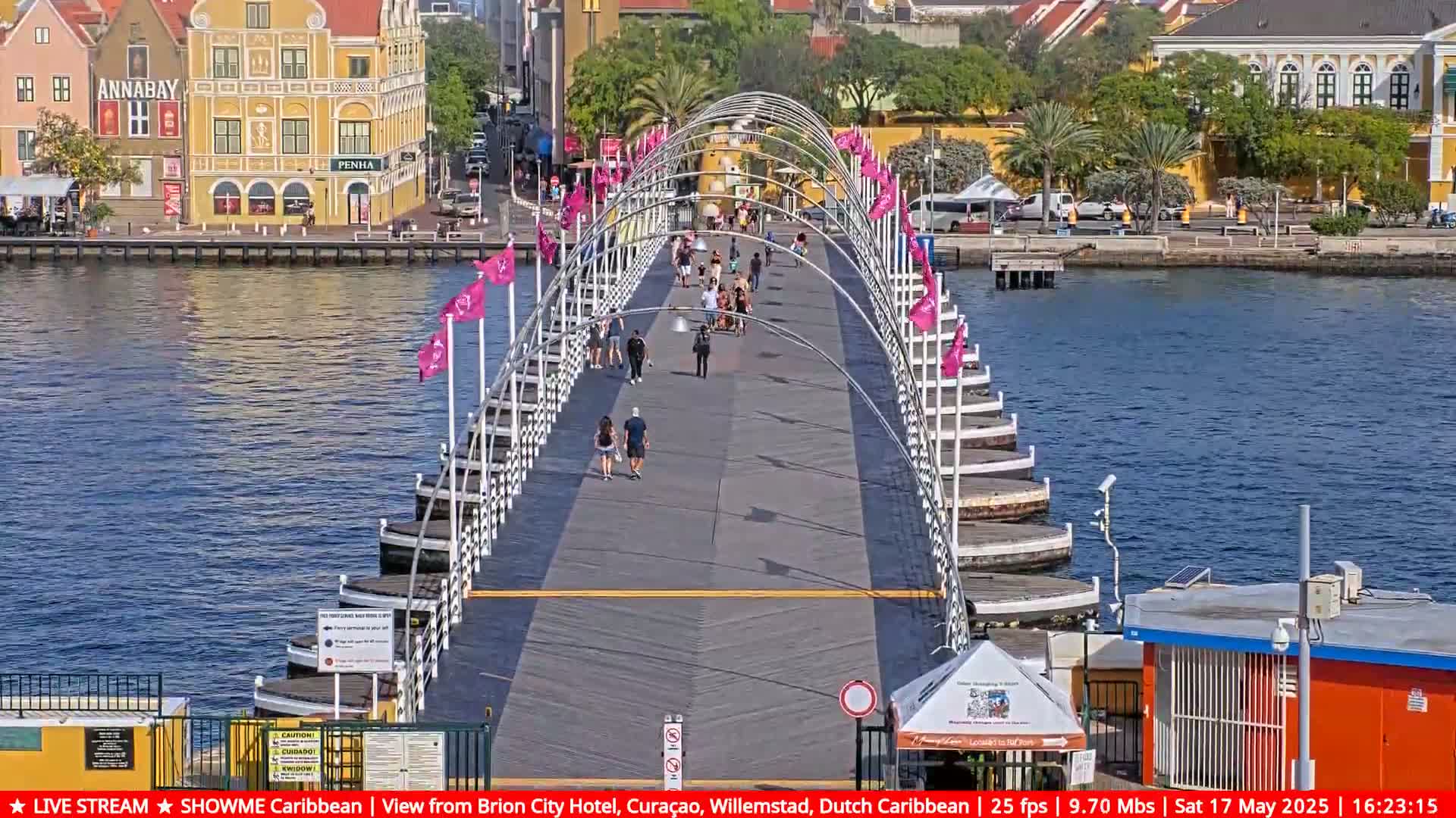 A pontoon bridge with pedestrians on it stretches across a calm body of water under a sunny sky, with colorful buildings visible in the background.