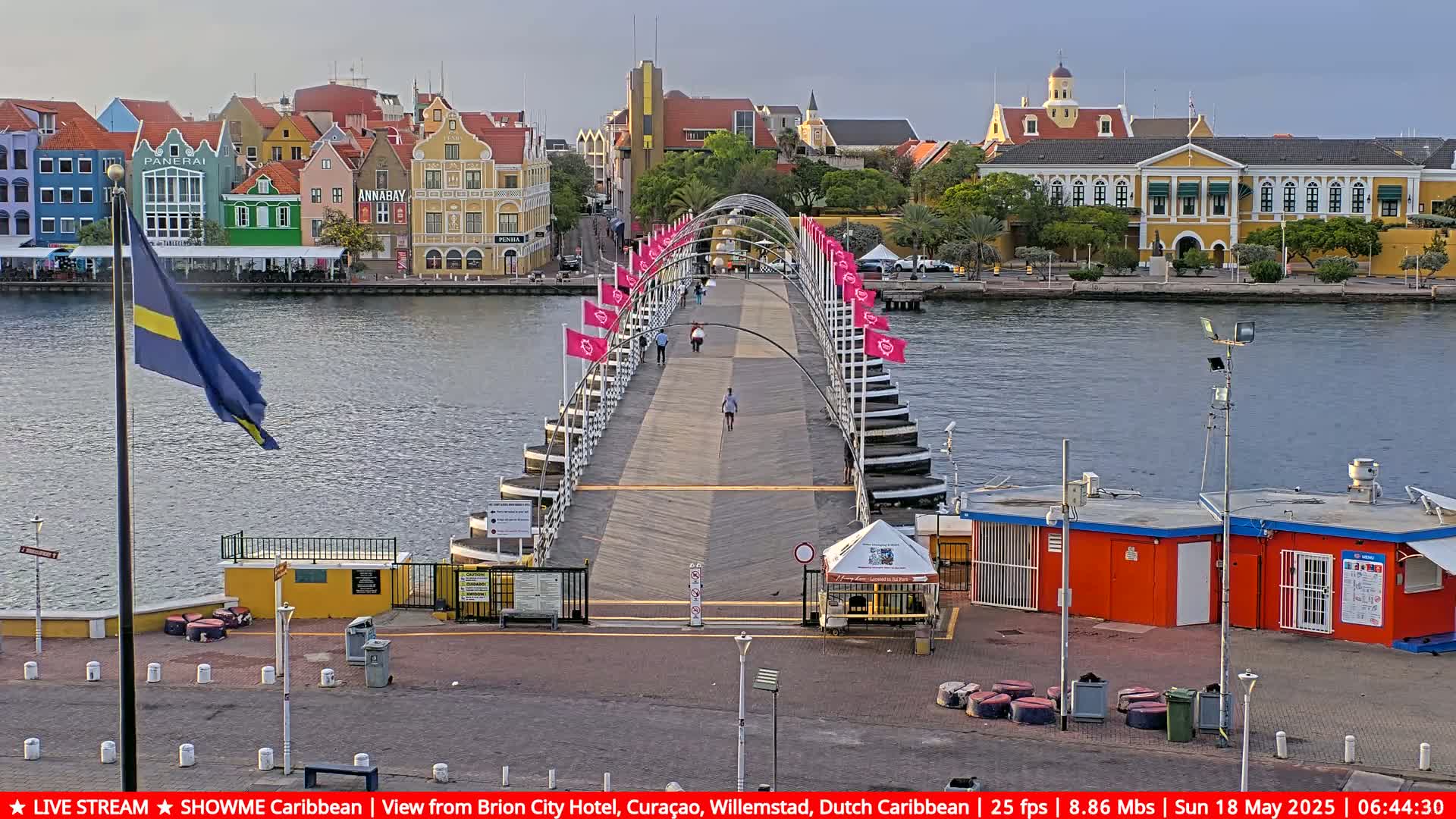 A long, arched pedestrian bridge stretches across a calm body of water toward colorful buildings under a partly cloudy sky.
