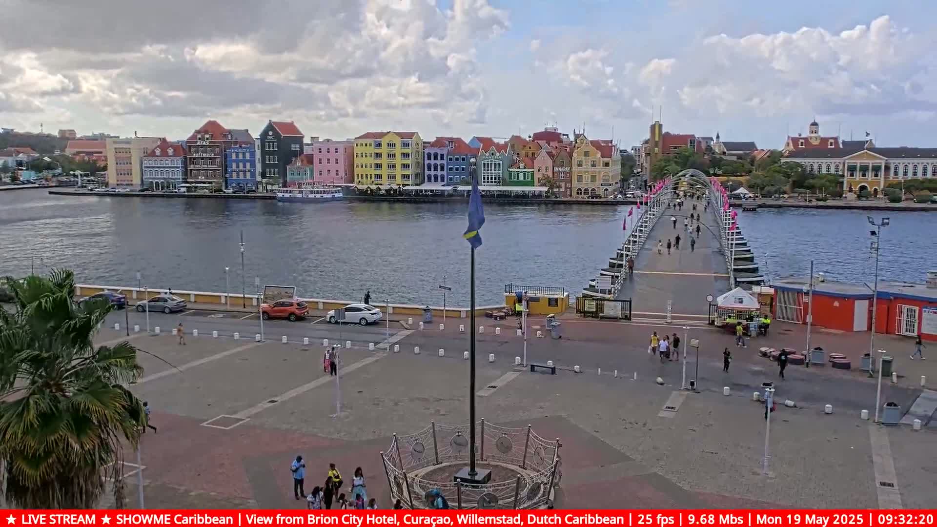 A partly cloudy day shows a colorful waterfront city with a pontoon bridge, several people walking on the bridge and plaza, and cars parked along the road.