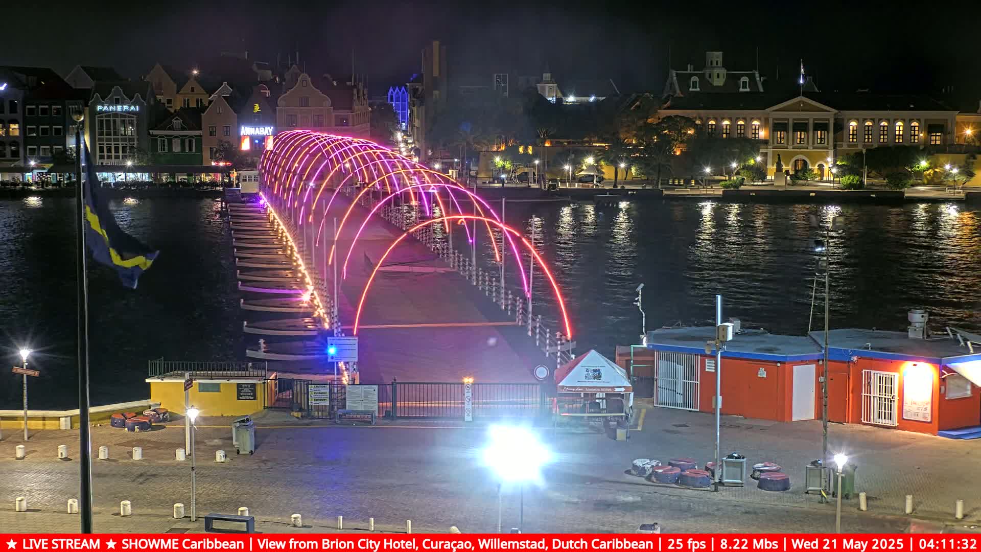 A brightly lit, arched pedestrian bridge spans a dark waterway at night, with buildings and a flagpole visible on the shore.