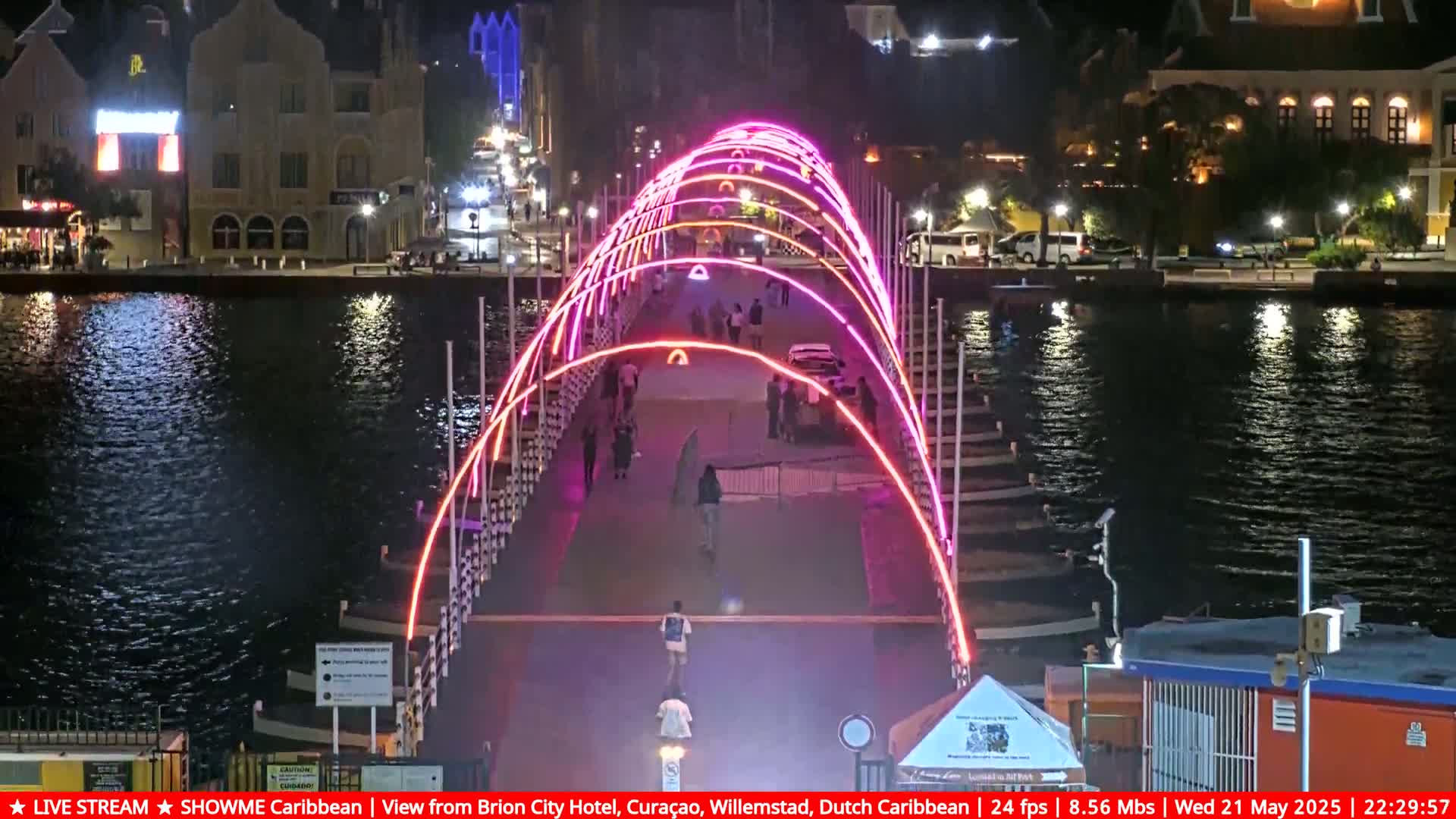 A brightly lit pedestrian bridge at night spans a calm waterway, with buildings and people visible on either side.