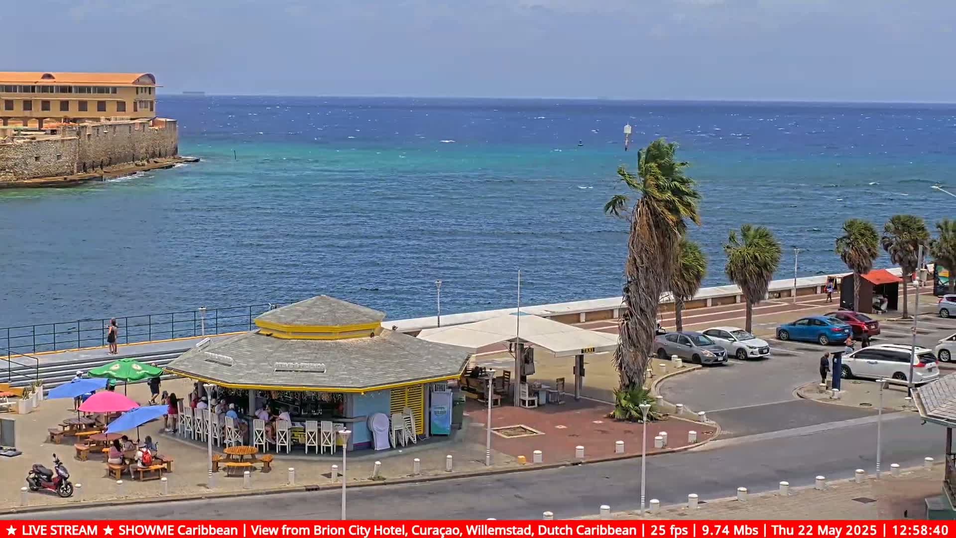 A waterfront scene on a sunny day shows a beach bar with patrons, palm trees, parked cars, and a stone building in the distance overlooking a calm, teal ocean.