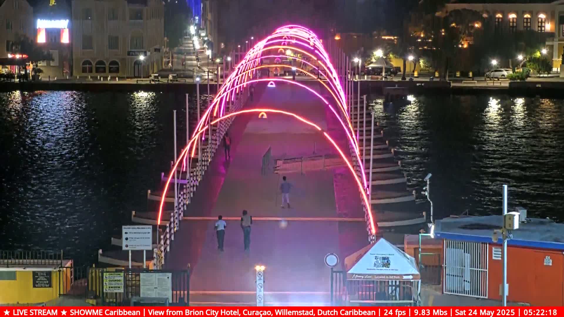 A lighted pedestrian bridge at night spans a calm waterway, with a few people walking across it.
