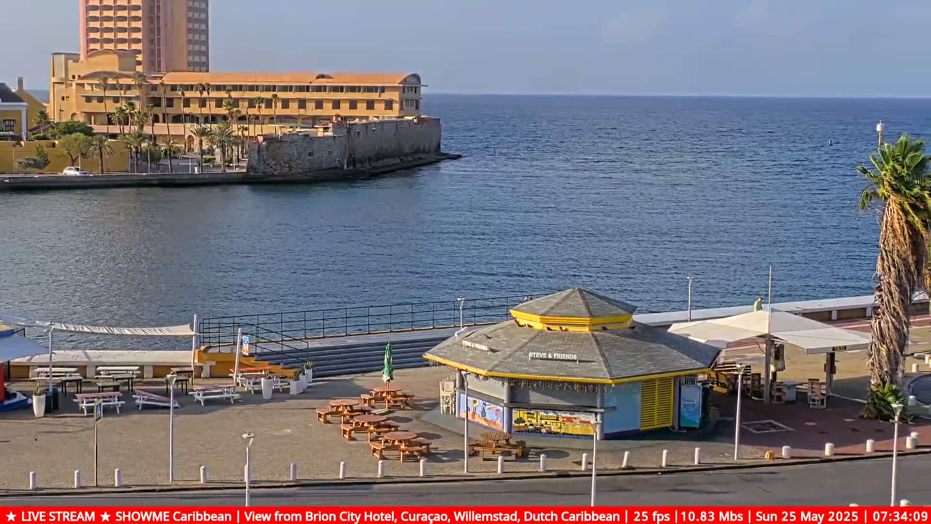 A waterfront scene on a sunny day shows a yellow kiosk with outdoor seating next to a calm body of water with a large building and palm trees in the background.