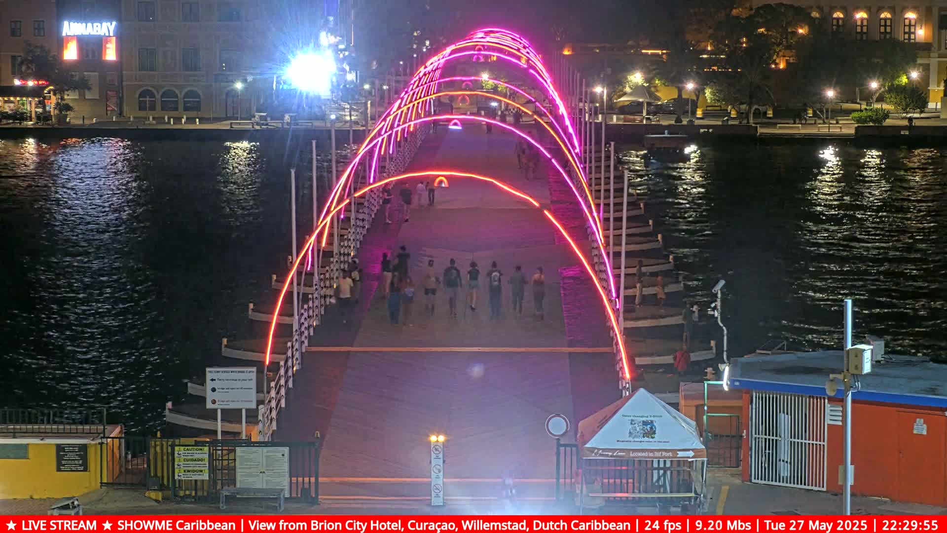 A brightly lit pedestrian bridge at night, arched with pink and orange neon lights, spans a dark waterway with people walking across it.