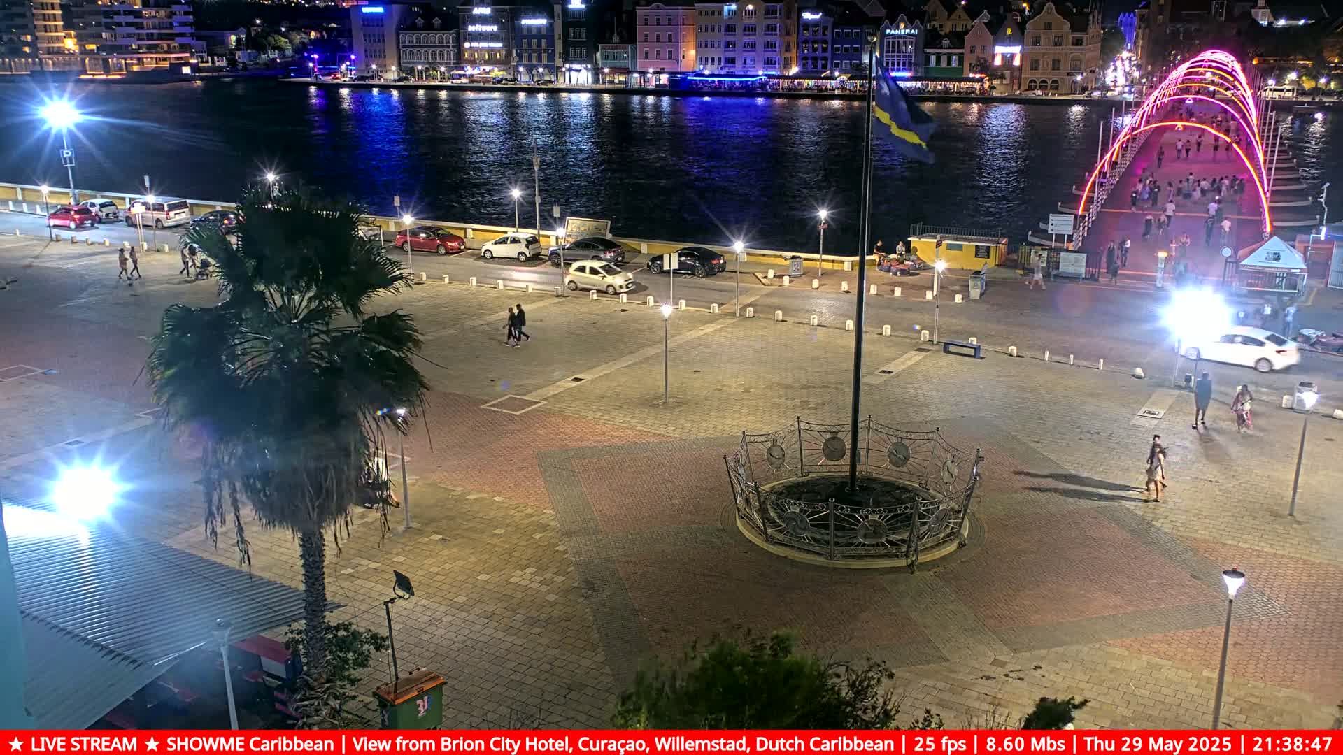 A nighttime view of a city square with a few people walking, cars parked nearby, a palm tree, and a brightly lit bridge in the background over a dark body of water.