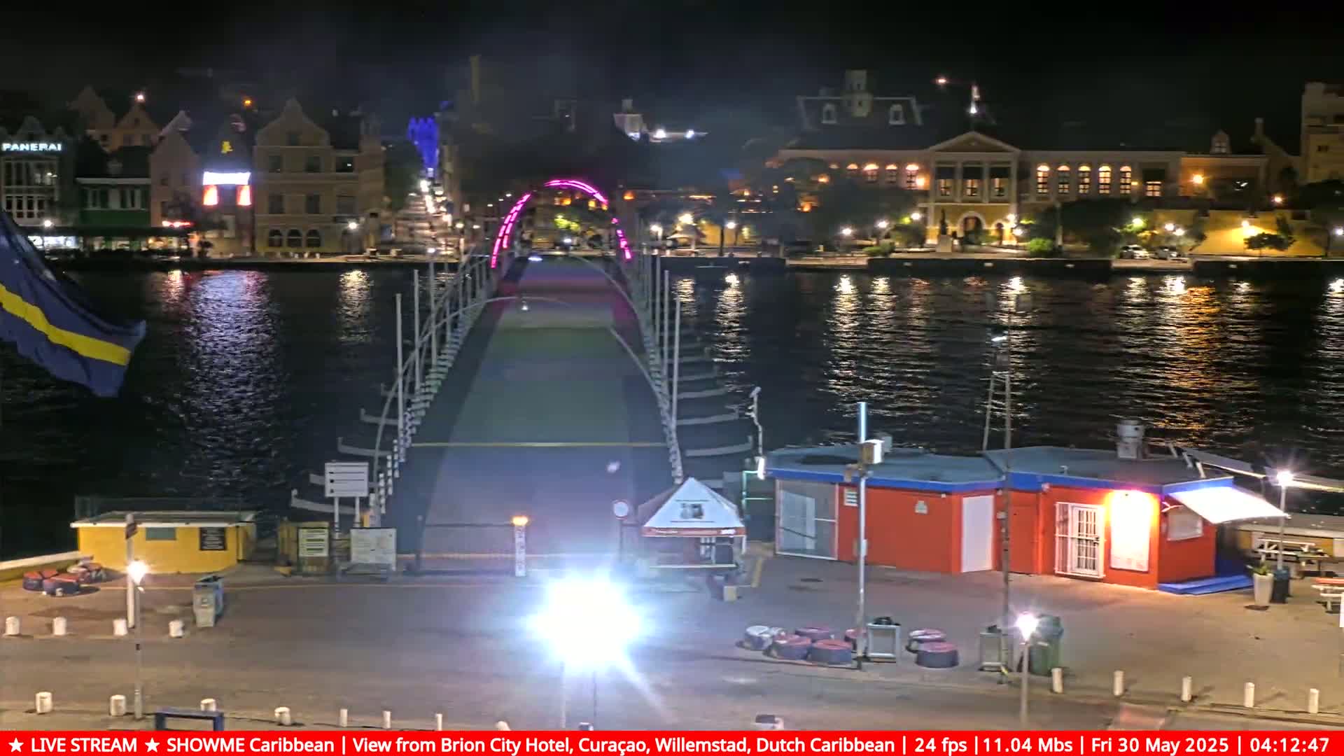 A nighttime view of a lighted pedestrian bridge spanning a calm body of water, with colorful buildings lining the waterfront.