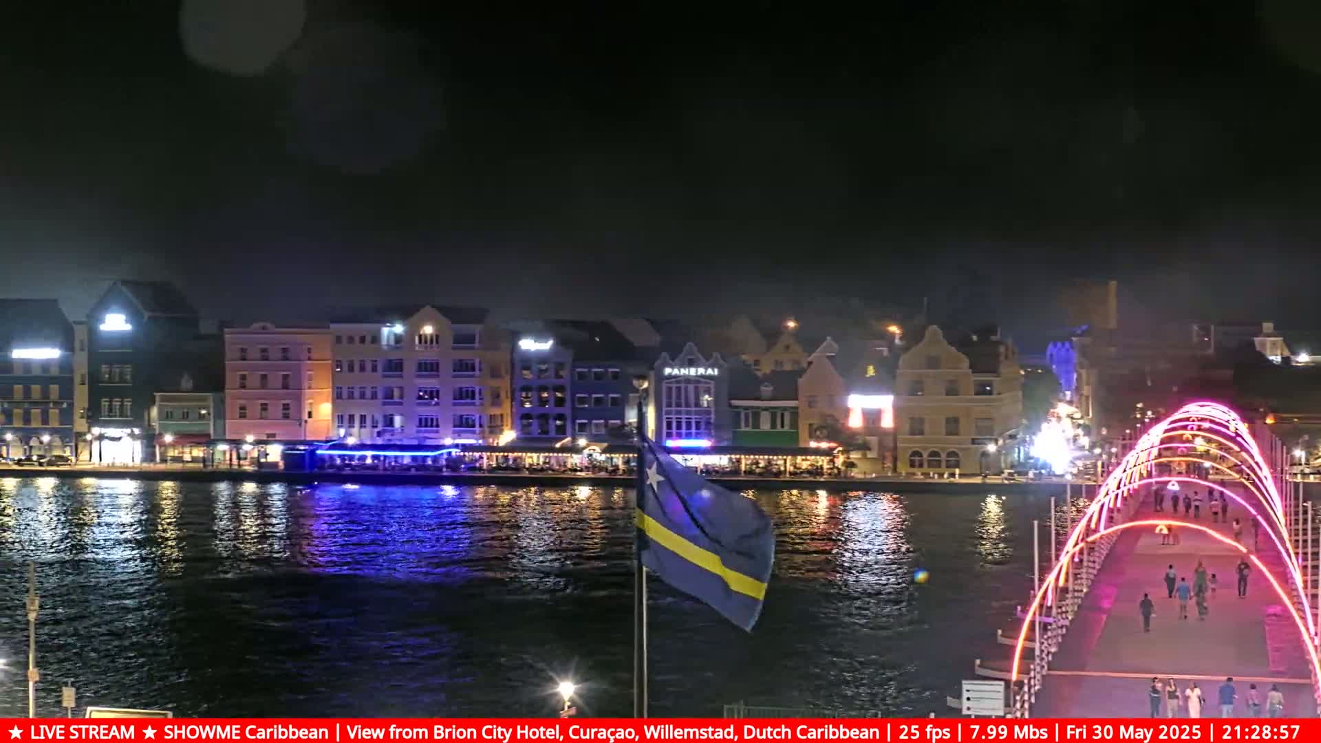 A nighttime waterfront scene shows brightly lit buildings along a calm body of water, with a bridge illuminated in pink and people walking across it.