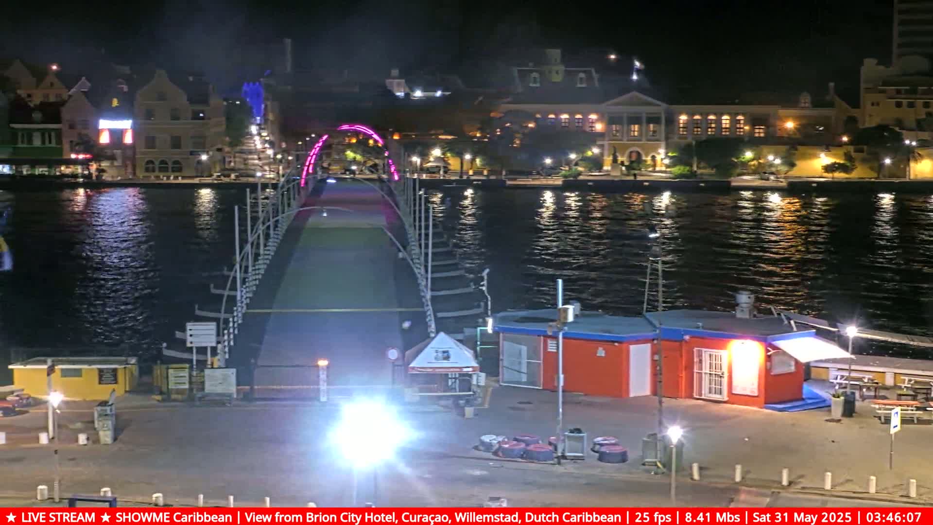 A pink-lit bridge spans a dark waterway at night, with buildings and small structures visible on both sides.
