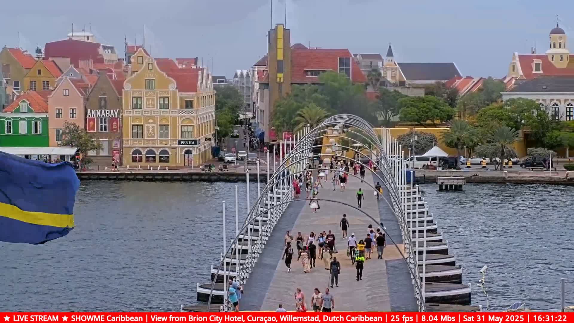 A pedestrian bridge over a waterway, flanked by colorful buildings, with many people walking across it under an overcast sky.